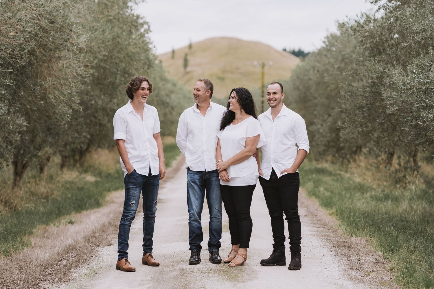 Four people wearing white shirts pose together on a dirt road surrounded by olive trees.
