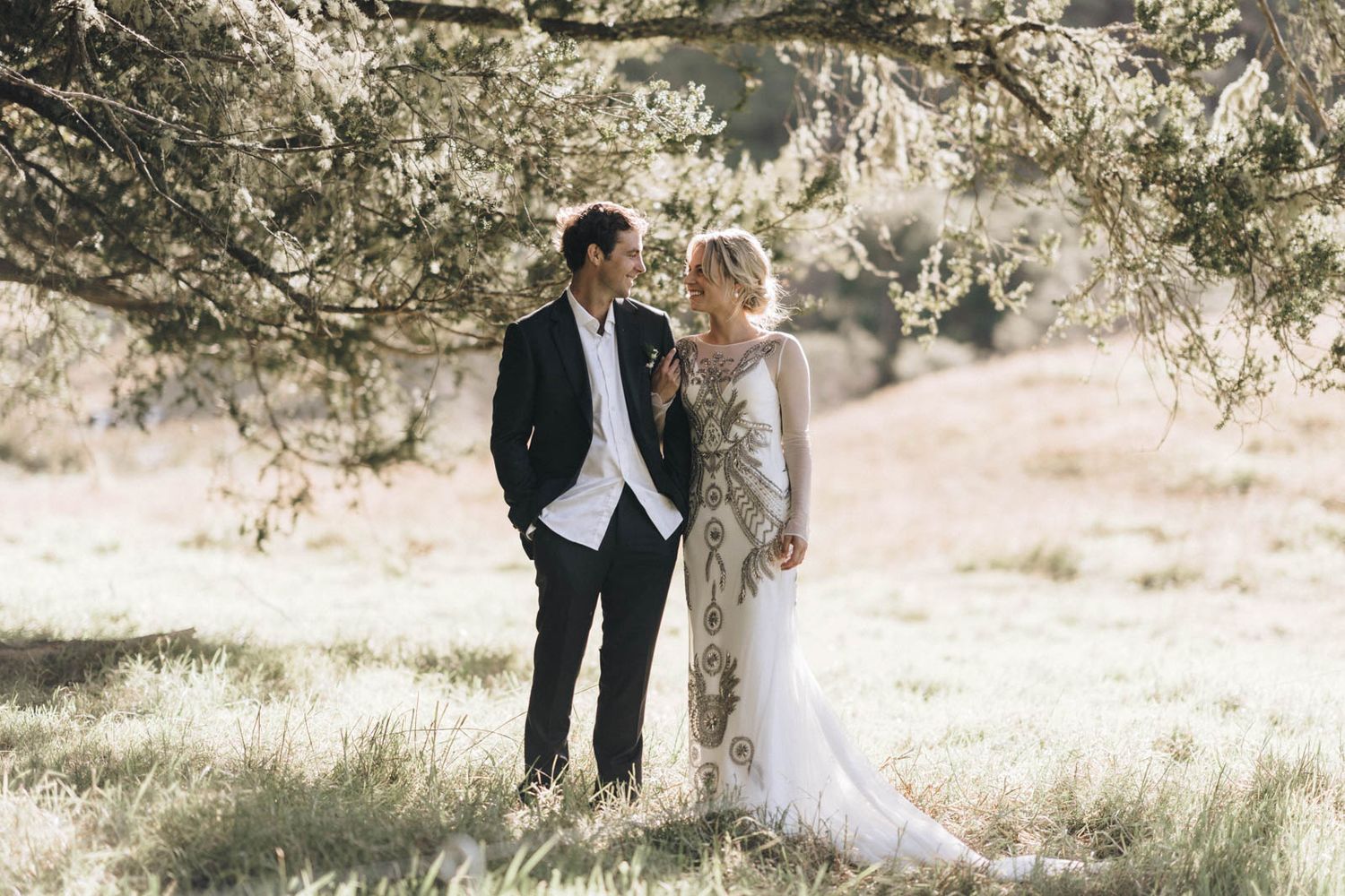 A wedding couple stands together in a sun-drenched meadow under a sprawling tree.
