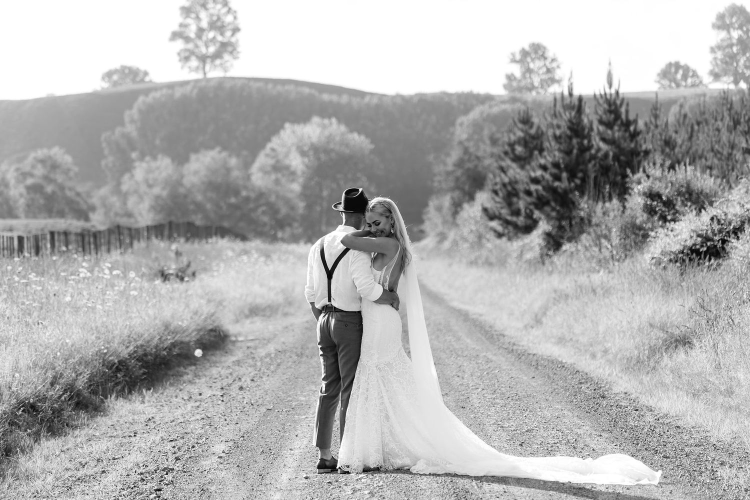 A bride and groom embrace on a scenic country road surrounded by rolling hills and trees in black and white.