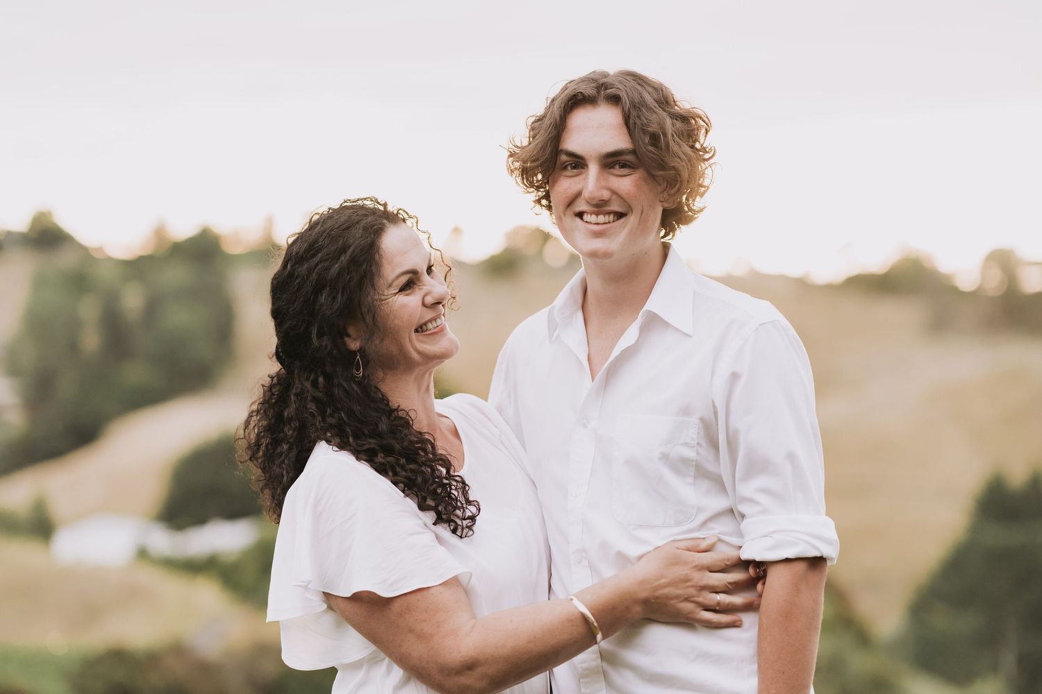 A couple in white clothing smiles and embraces while standing together in a sunlit outdoor setting.