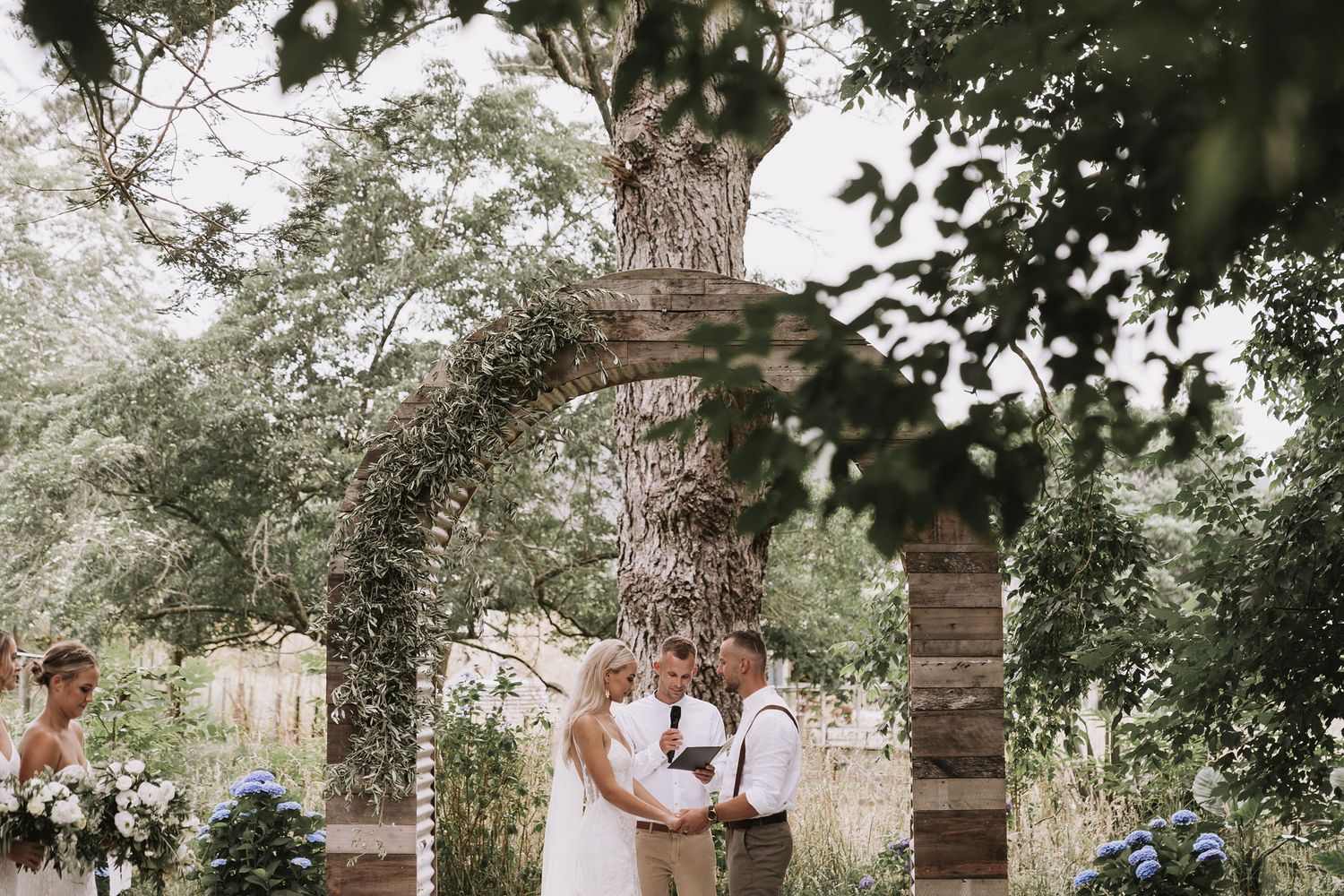 A romantic outdoor ceremony takes place under a rustic stone arch adorned with greenery in a garden setting.
