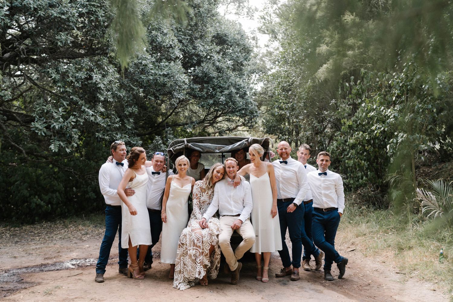 Wedding party wearing white and navy outfits poses together on wooded dirt path for outdoor group photo.