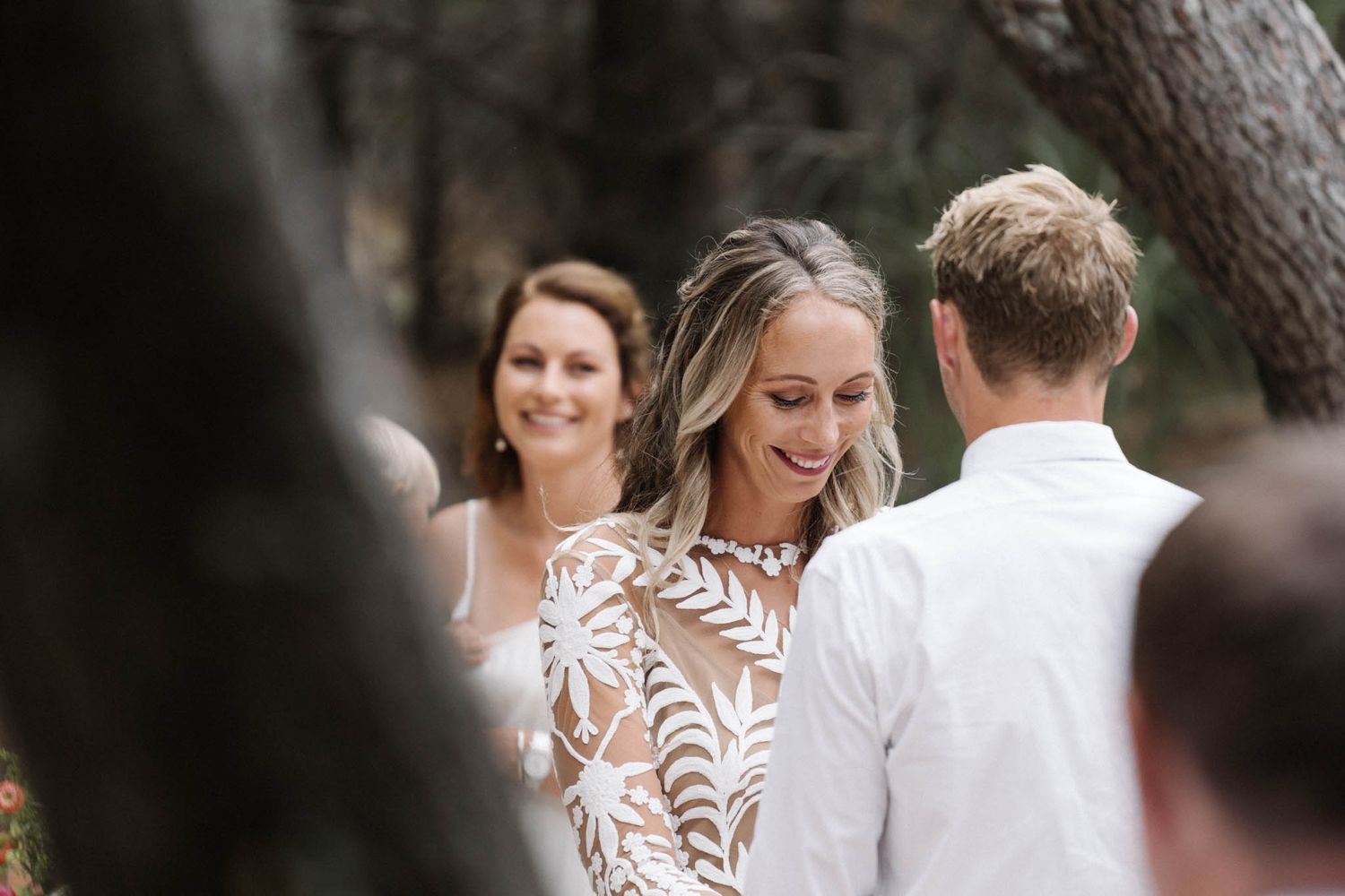 A bride in an intricate lace dress exchanges vows during an intimate outdoor forest wedding ceremony.