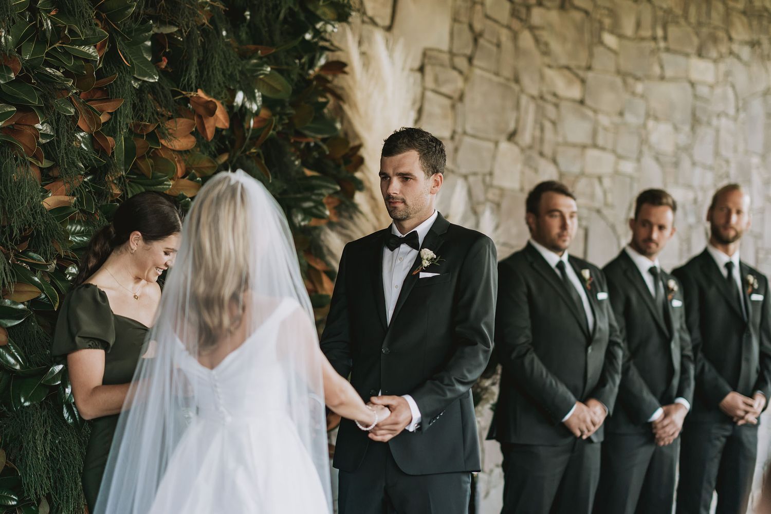Elegant wedding ceremony scene with bridal party in black suits and veiled bride against stone wall with greenery.