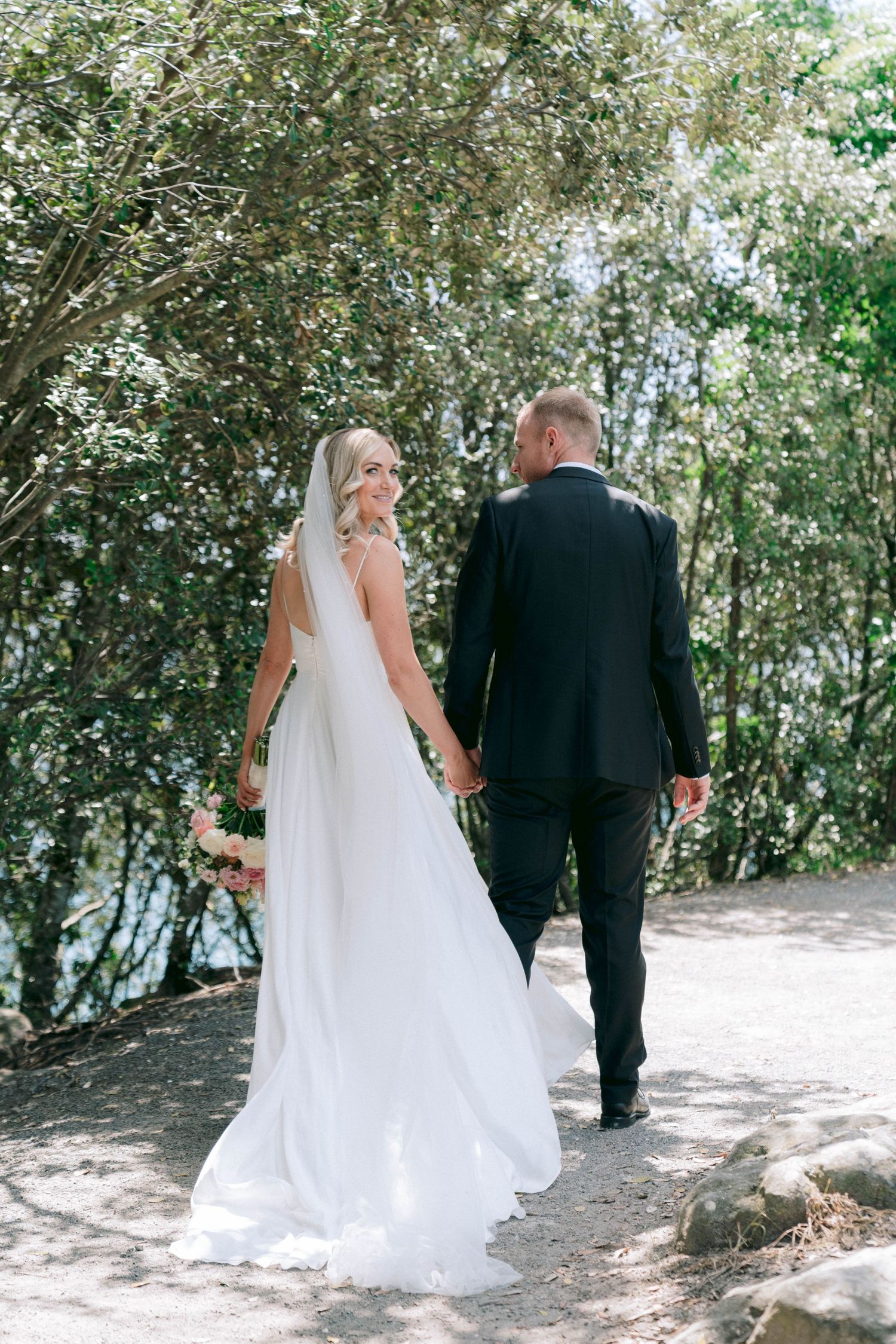 Bride and groom walking together through a tree-lined pathway in formal attire.
