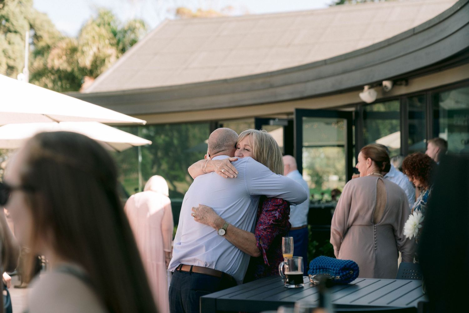 Wedding guests embrace during outdoor reception with building and umbrellas in background.