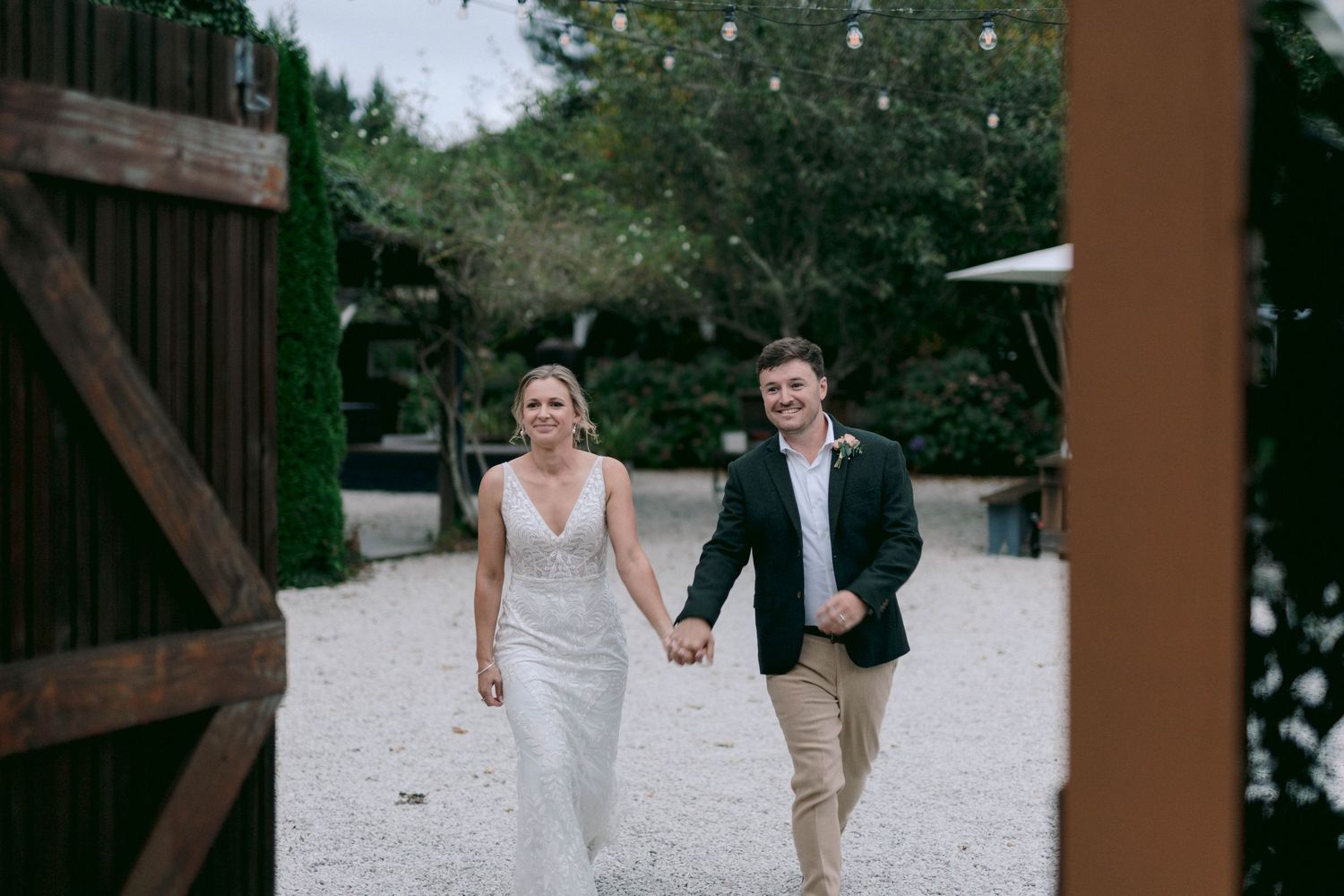 Couple walking hand in hand along a gravel path near wooden barn doors.