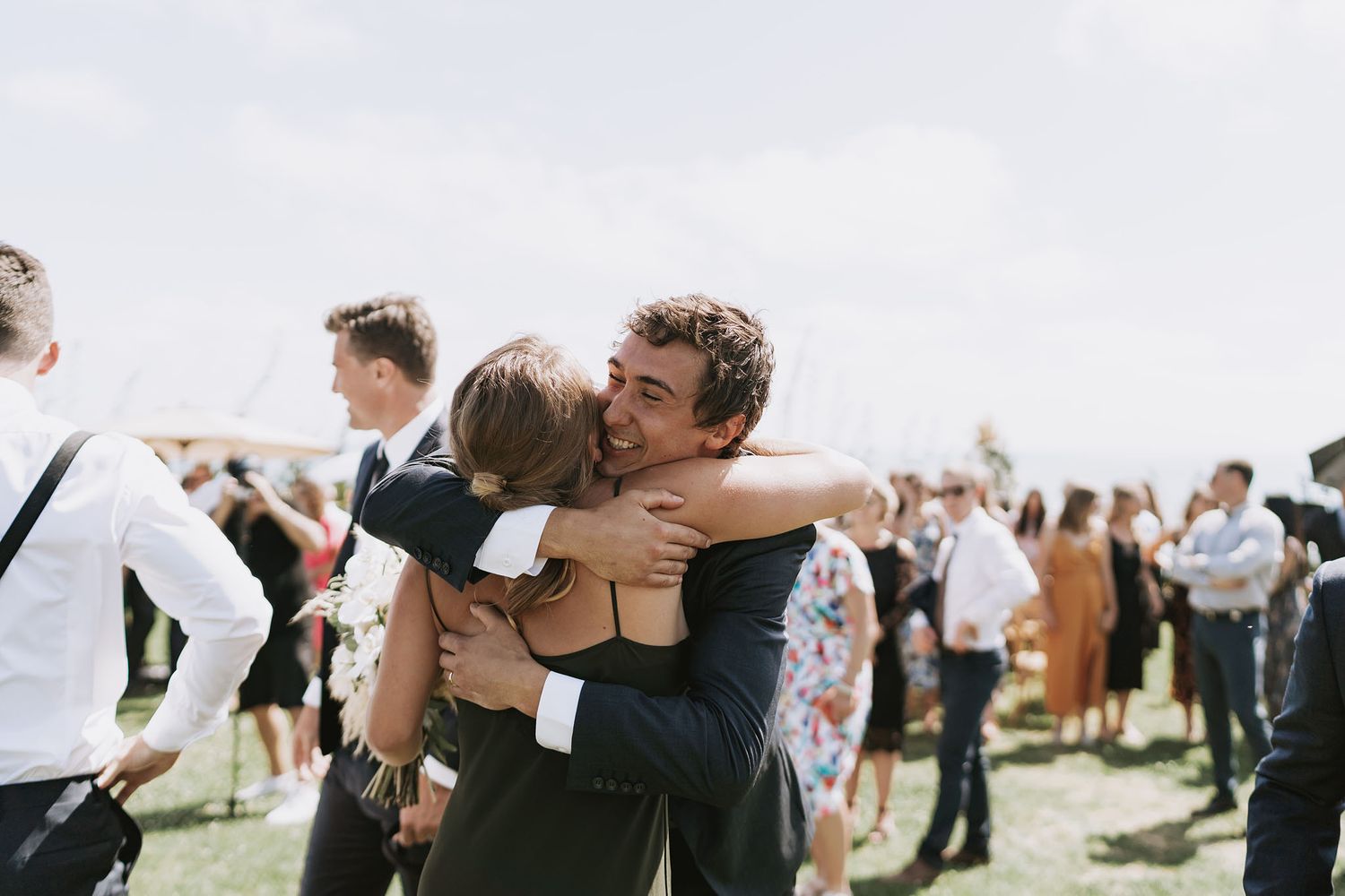 Wedding guests embrace each other during an outdoor celebration with other attendees visible in the background.
