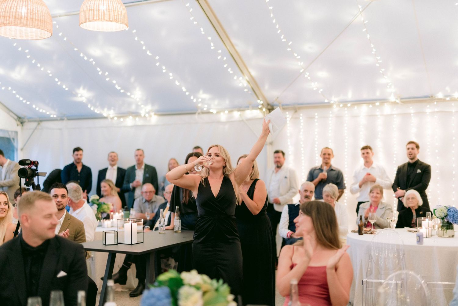 Wedding guests celebrate under a tent decorated with twinkling lights during an evening reception.