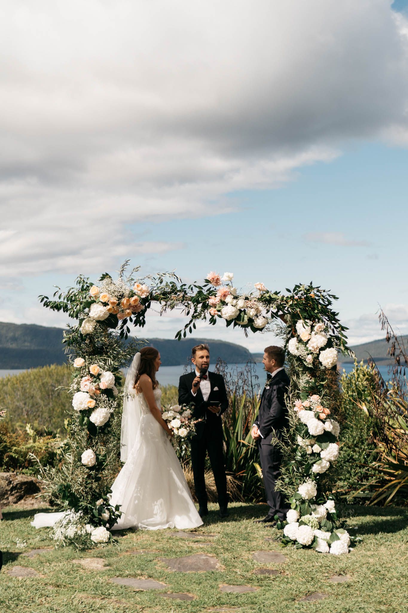 An elegant floral wedding arch with white roses and greenery stands on a cliffside overlooking a scenic mountain landscape.
