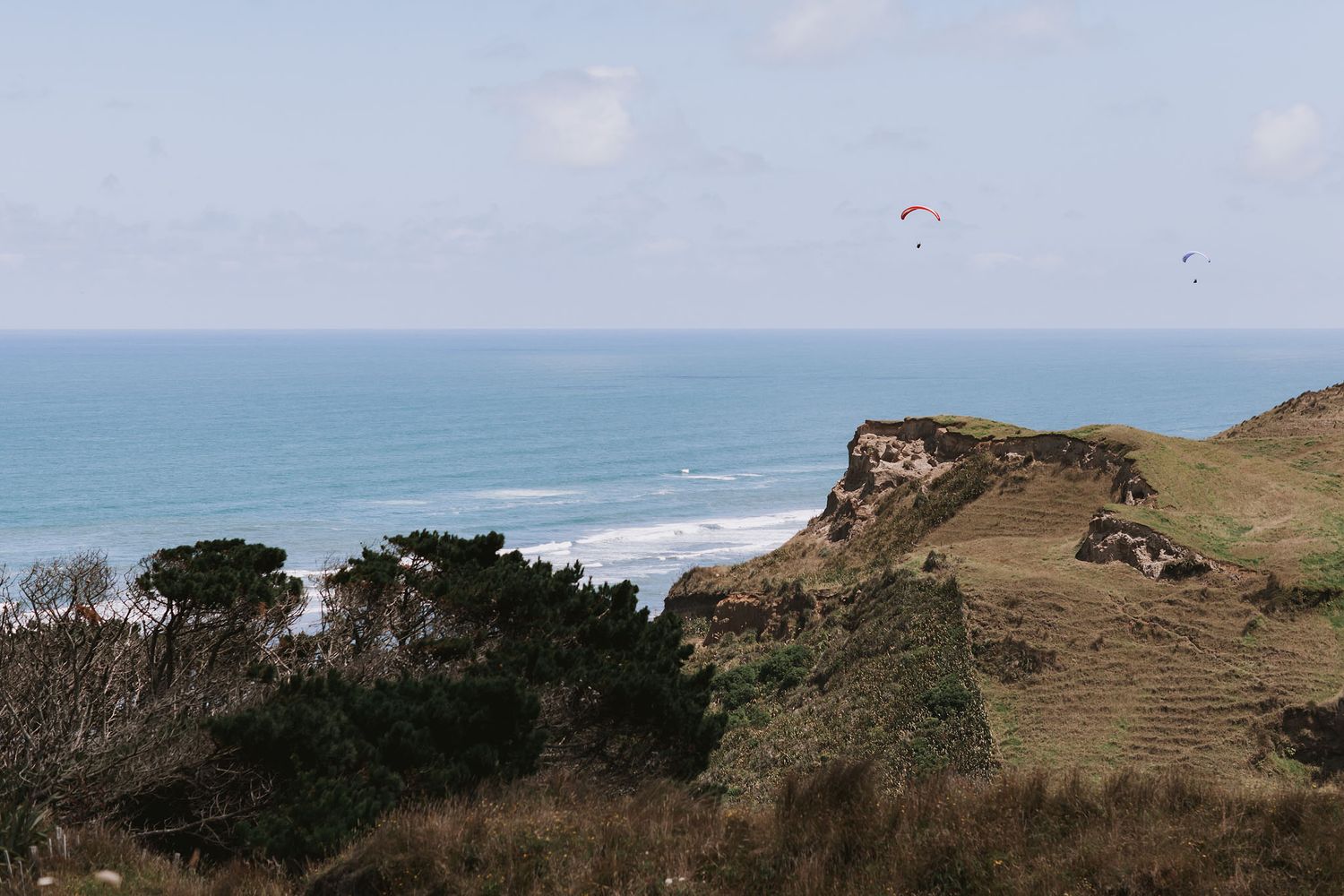 Paragliders soar above rugged coastal cliffs overlooking the blue ocean on a cloudy day.