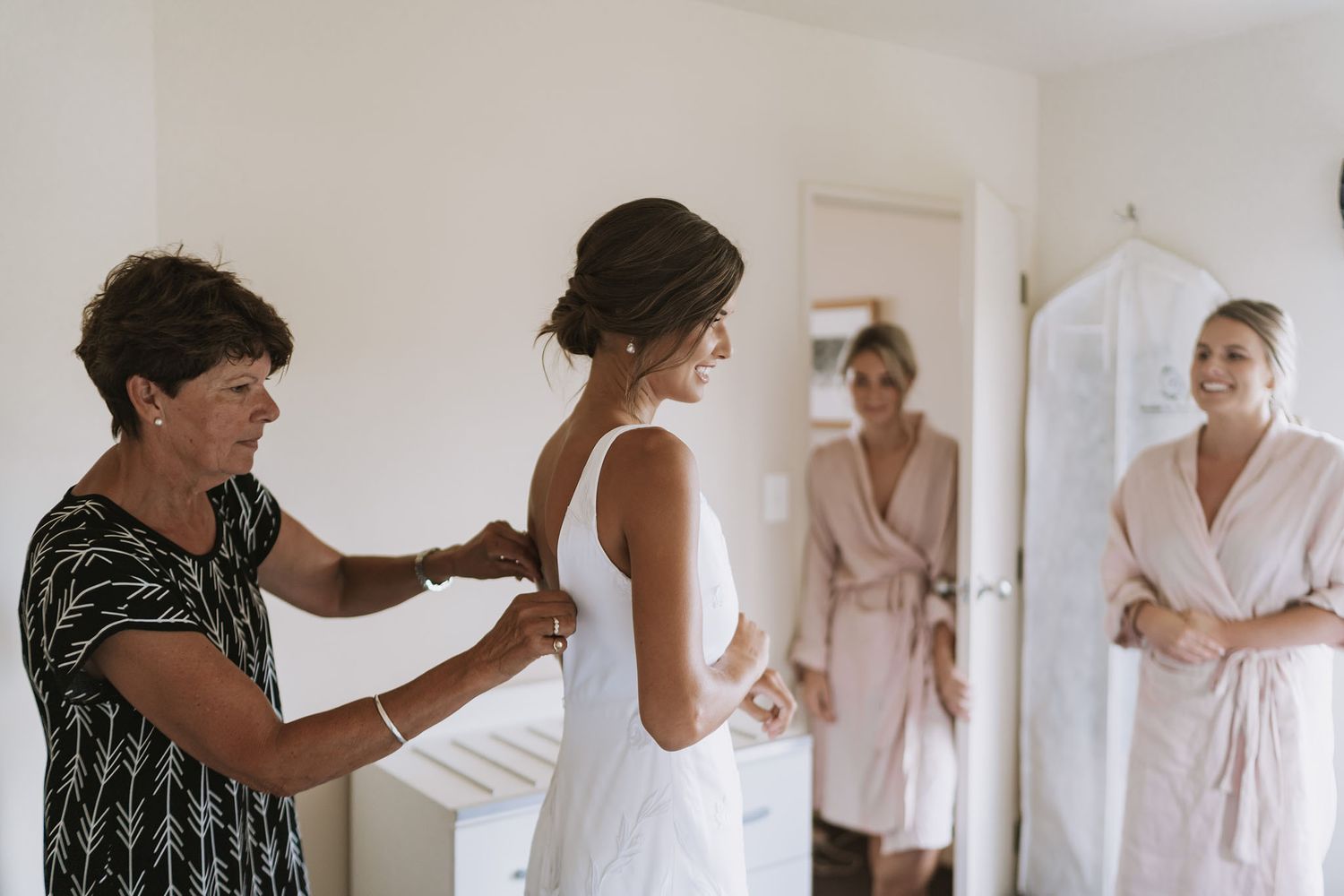 A person helps adjust a white wedding dress while two others in robes watch during wedding preparations.