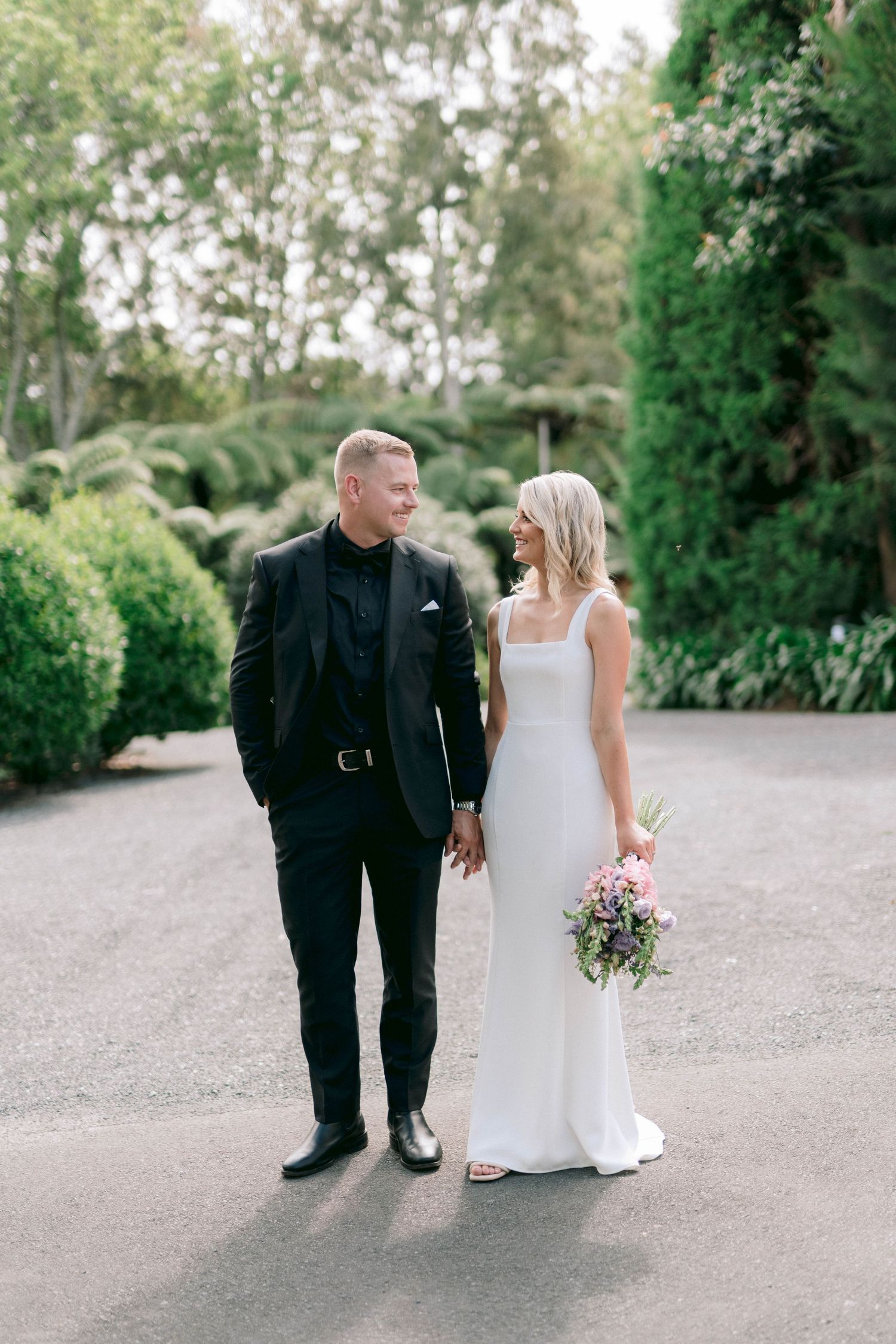 A couple in wedding attire smiles and walks together on a gravel path surrounded by lush greenery at an outdoor venue.