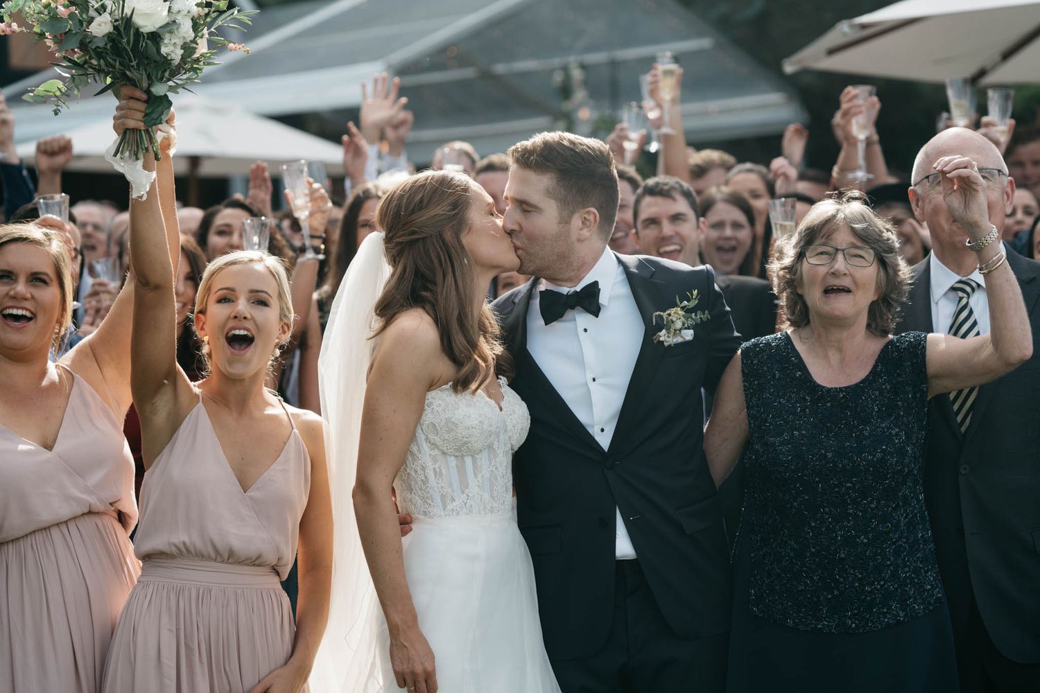 Wedding couple kisses during outdoor celebration as guests cheer enthusiastically in background.