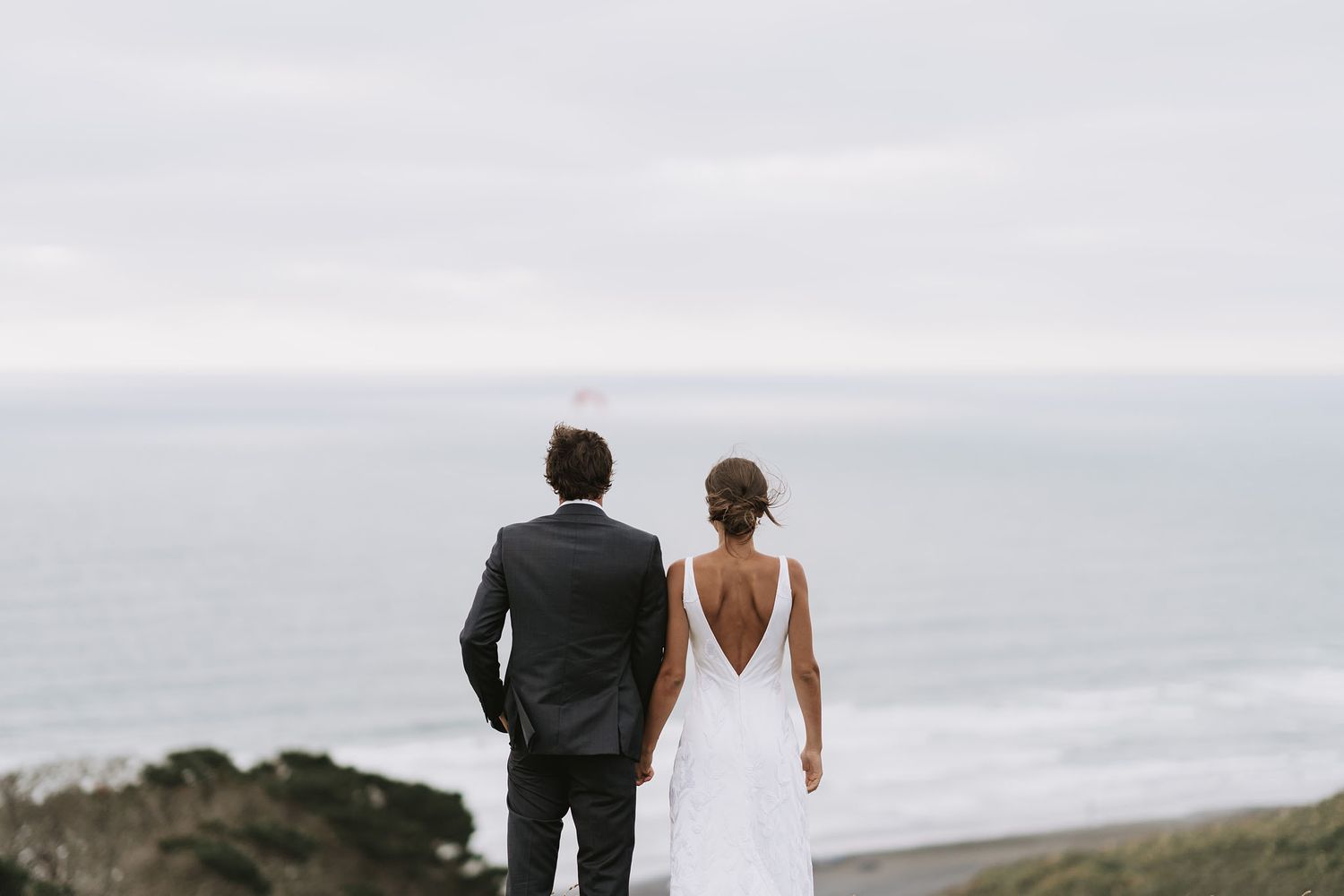 A couple in formal attire walks along the cliff edge overlooking the ocean during an intimate wedding ceremony.