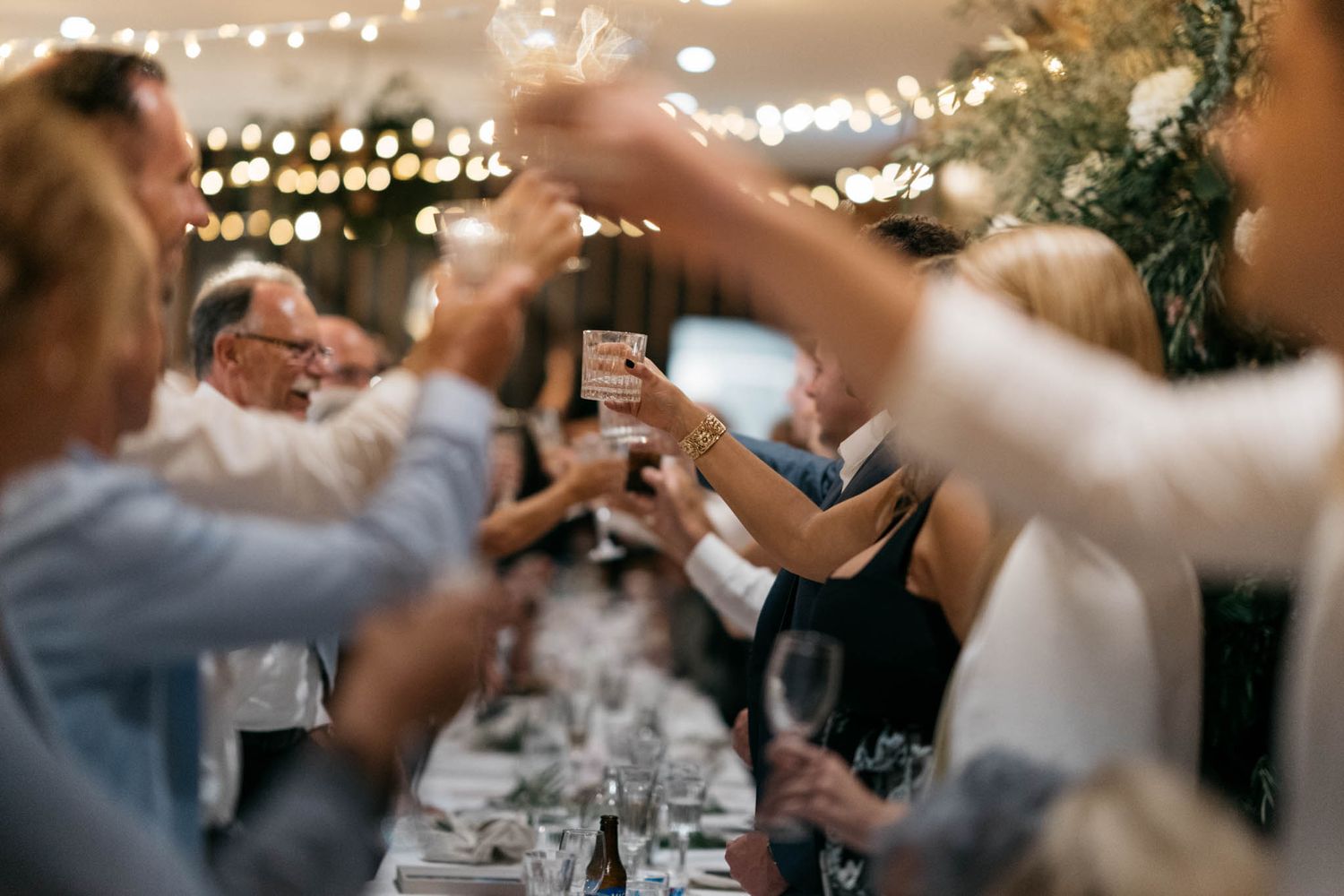 People raising wine glasses in a toast during an elegant dinner celebration with string lights overhead and table settings below.