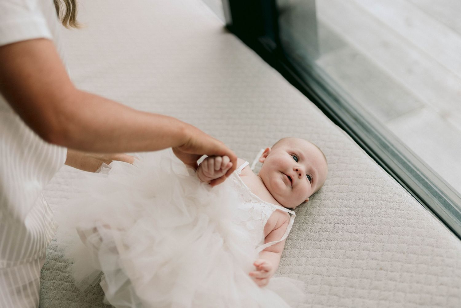 Baby wearing a white tulle dress lies on carpet beside window while being gently touched by parent's hand.