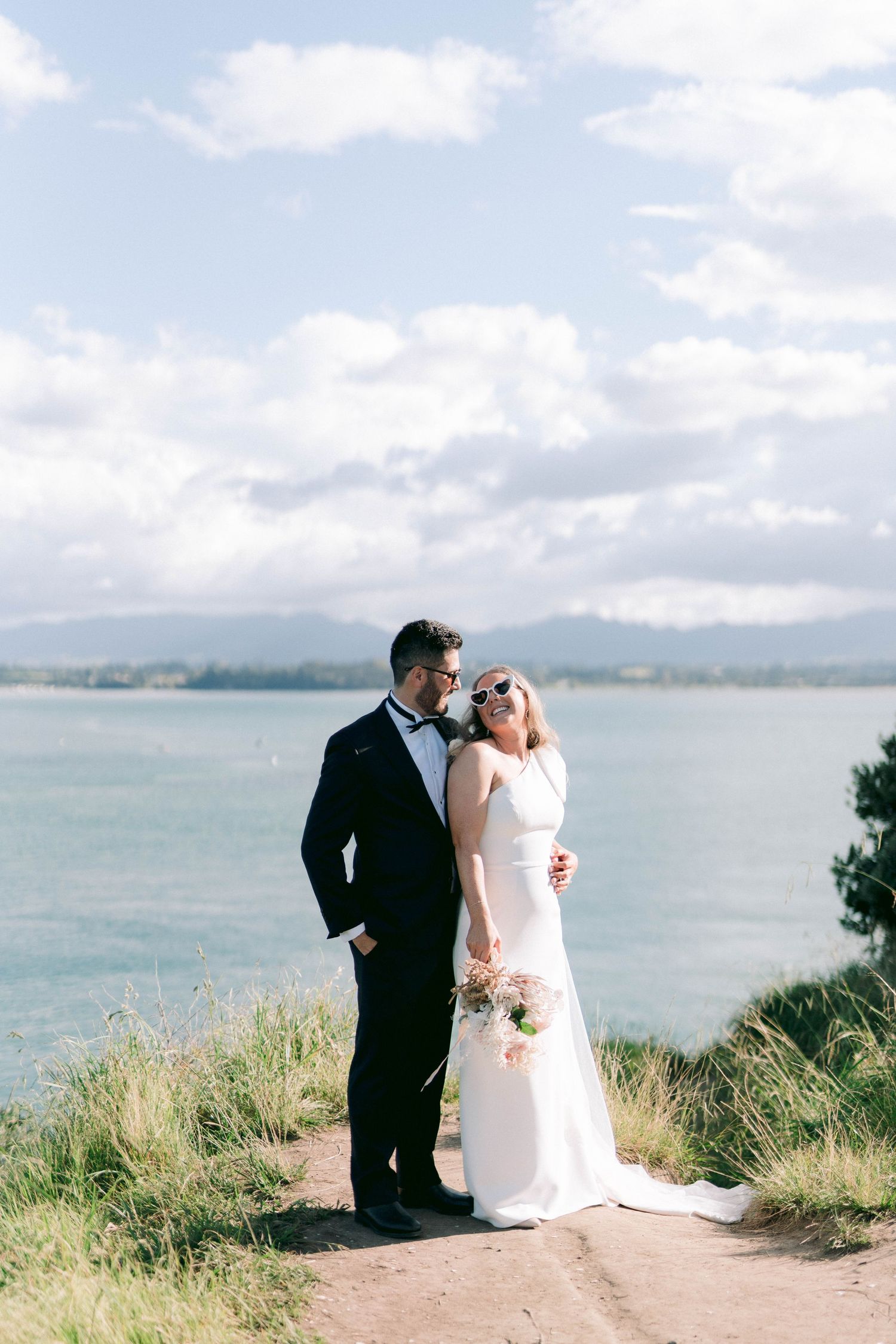 Newlywed couple sitting together on coastal cliff overlooking ocean with mountains in background.