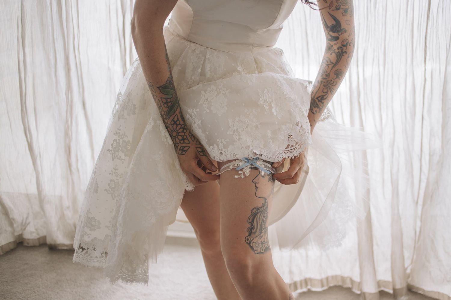 Person in white lace wedding dress shows leg garter and tattoos against sheer curtain backdrop in intimate boudoir setting.
