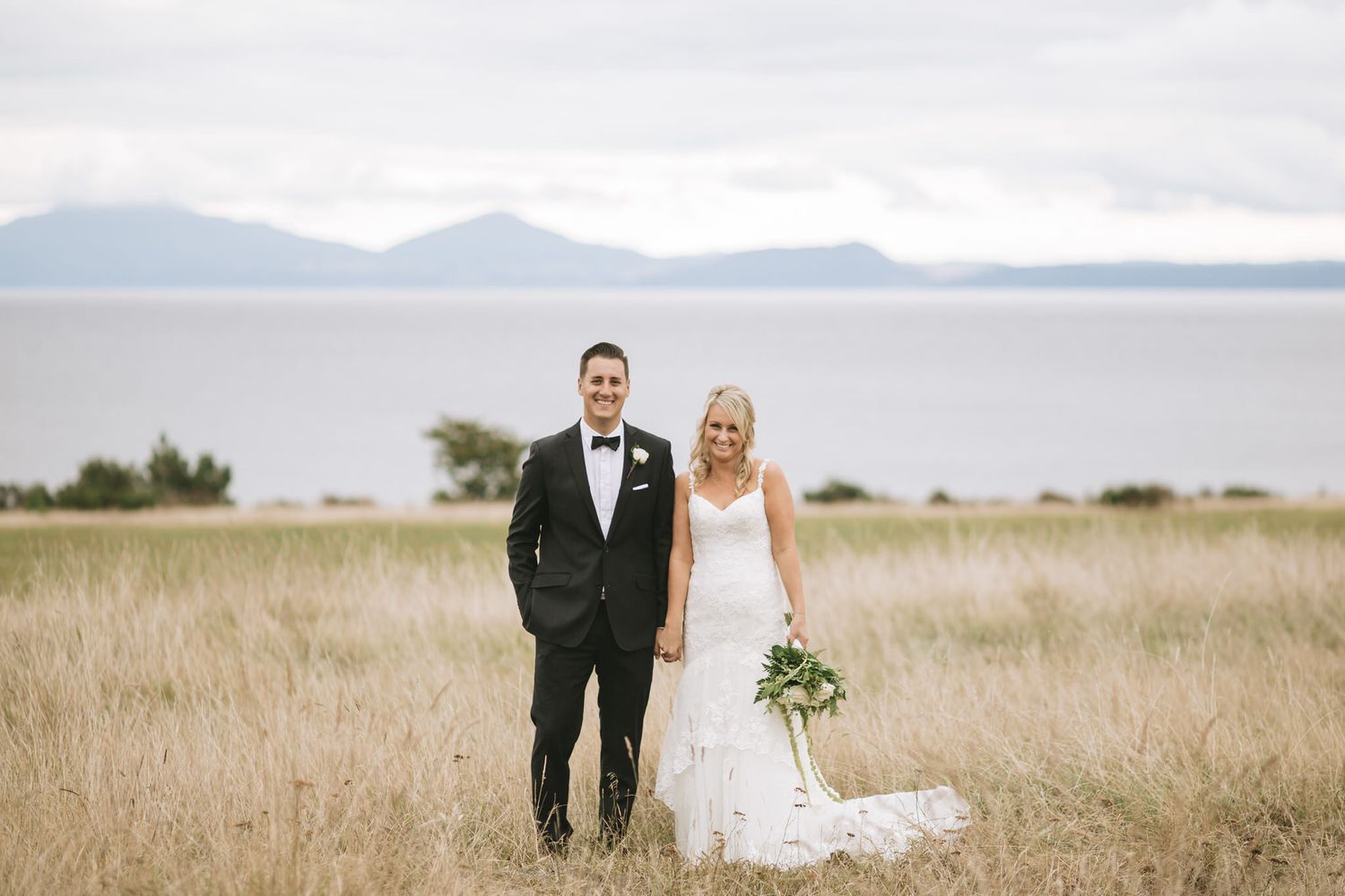 Couple stands together in formal attire on grassy bluff overlooking peaceful mountain lake on overcast day.