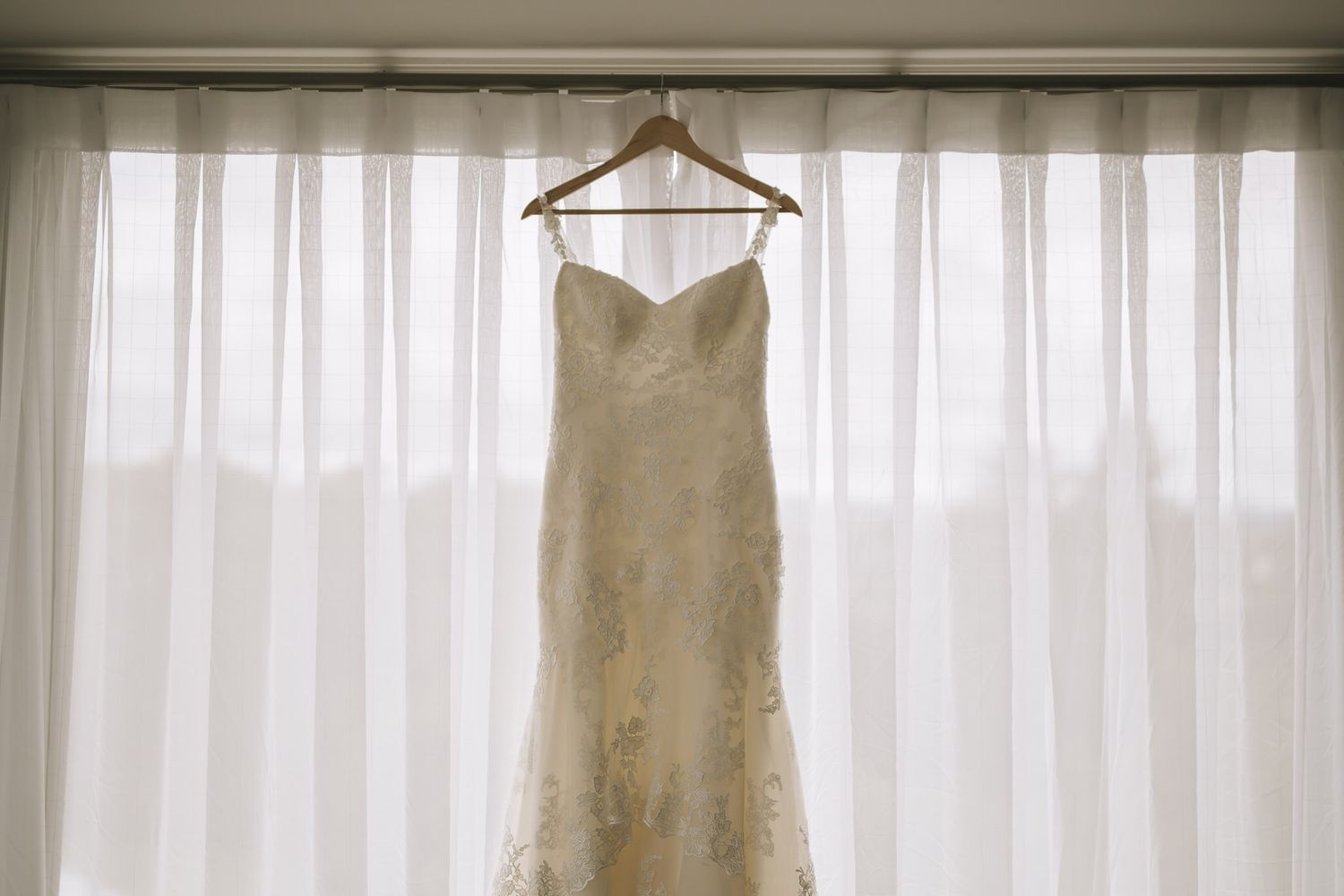 A white lace wedding dress hangs elegantly against sheer curtains in natural window light.