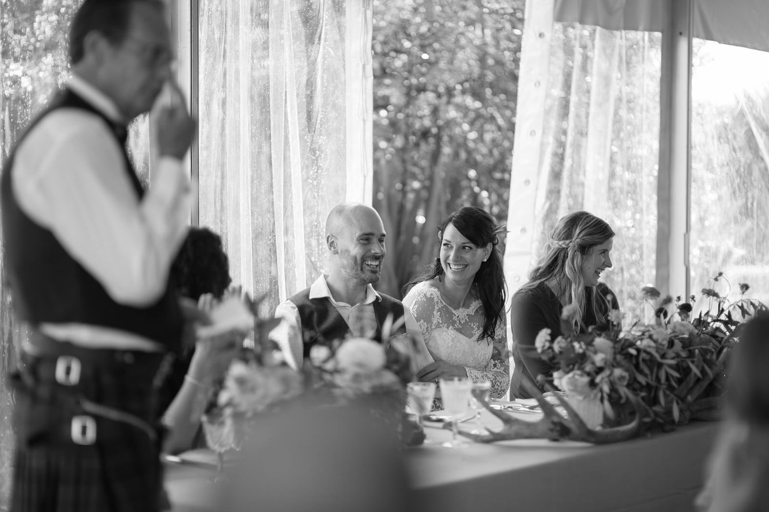 A couple shares a romantic kiss during their wedding ceremony by a window with soft light filtering through sheer curtains.