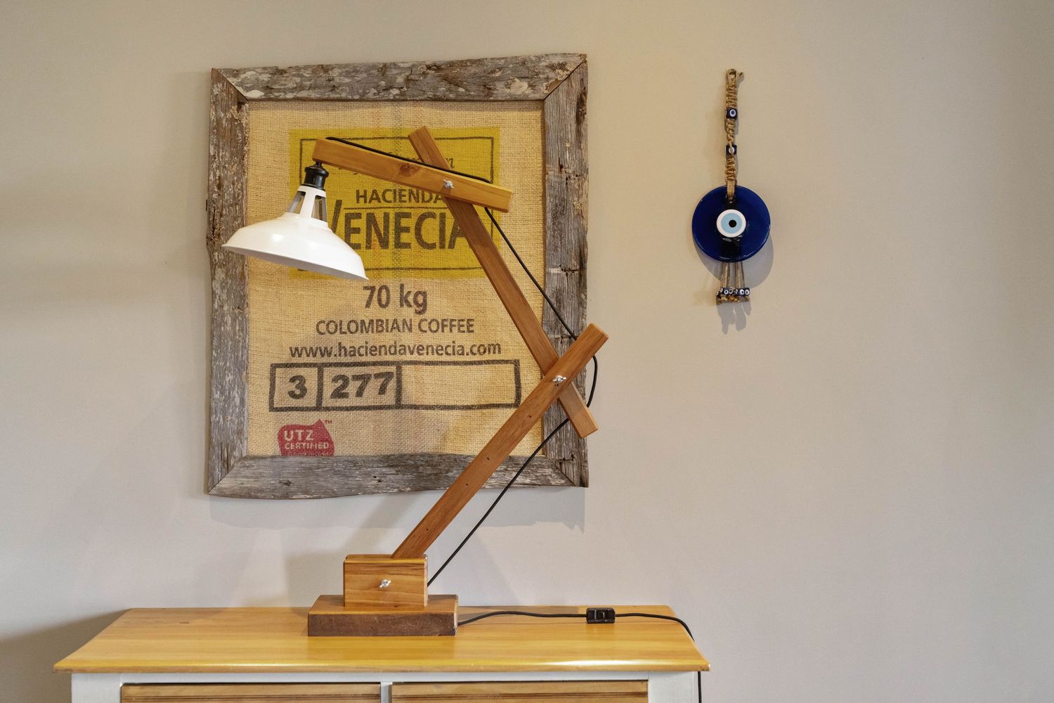 A wooden desk lamp and framed vintage poster on white wall with decorative blue evil eye charm hanging nearby.