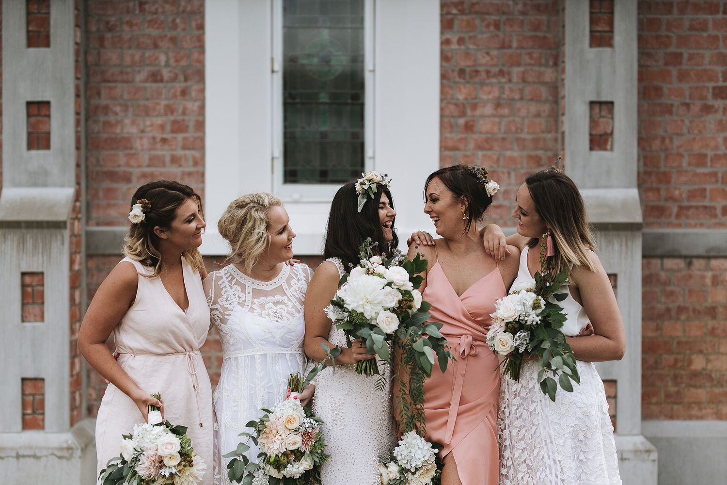 A row of bridesmaids in pink dresses holding white floral bouquets stand together on a brick building's steps.