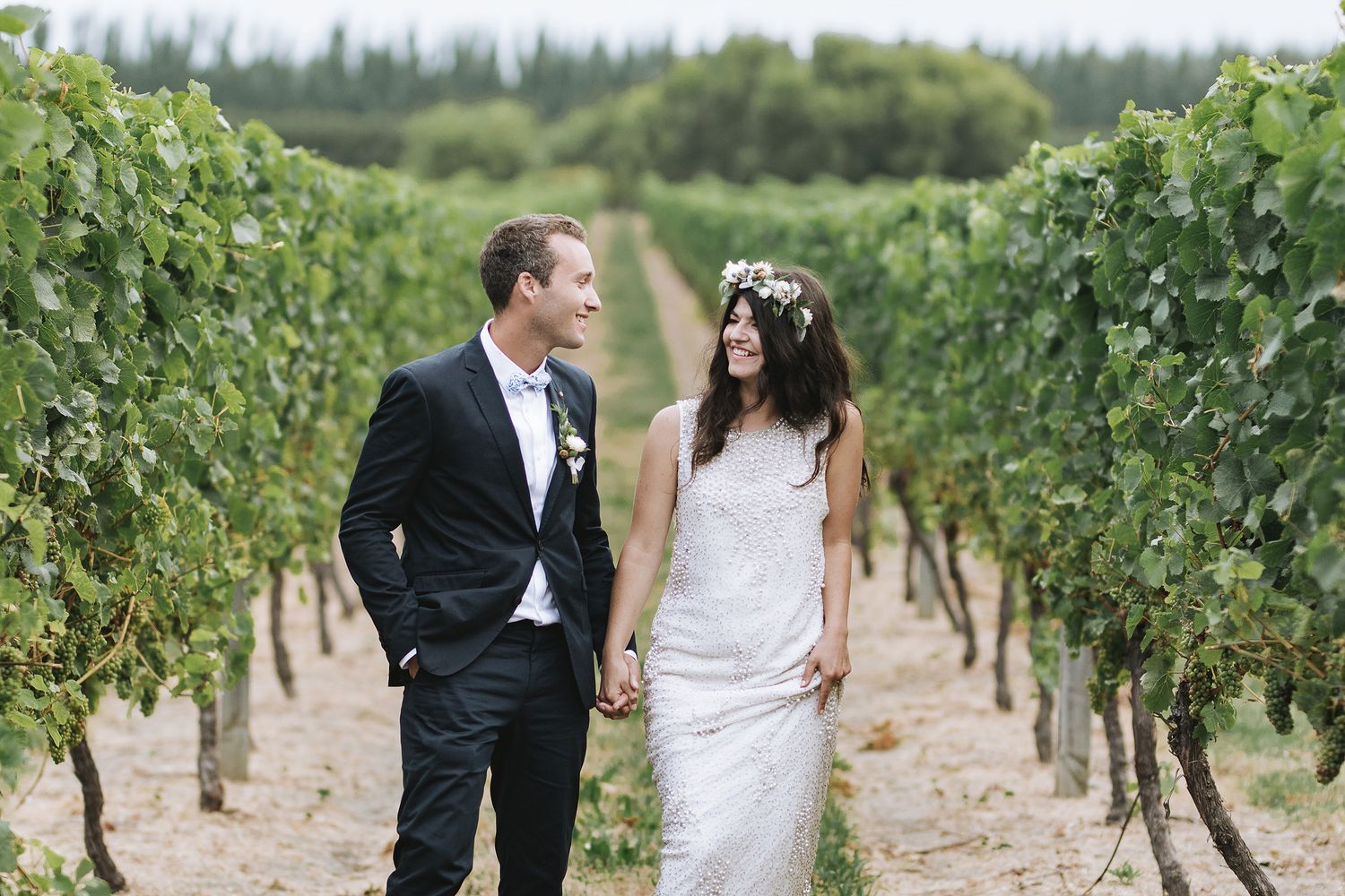 Happy couple in wedding attire walking through vineyard rows holding hands among green grapevines.