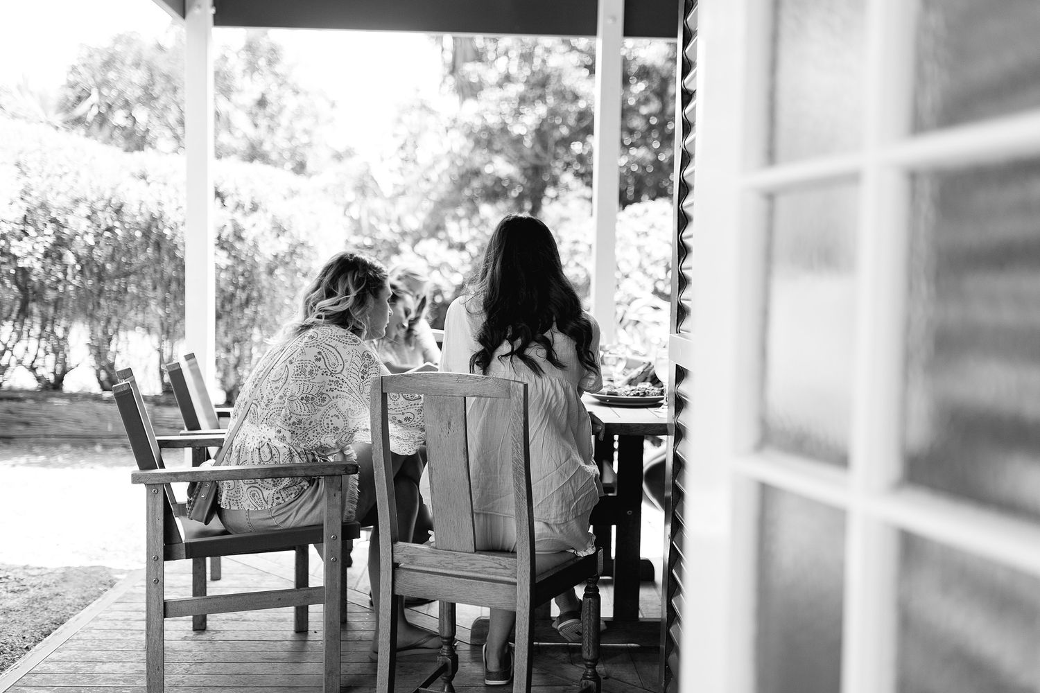 Two people sitting together in wooden chairs on a sunlit porch overlooking a garden.
