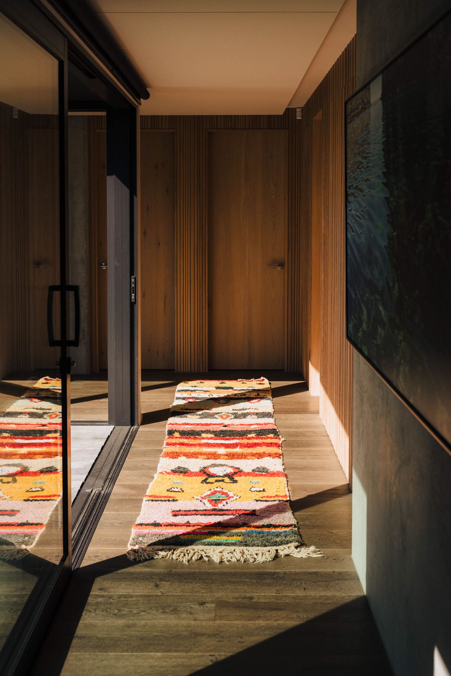 Sunlit hallway with wooden walls, hardwood floors, and colorful striped runner rugs leading to closed doors.