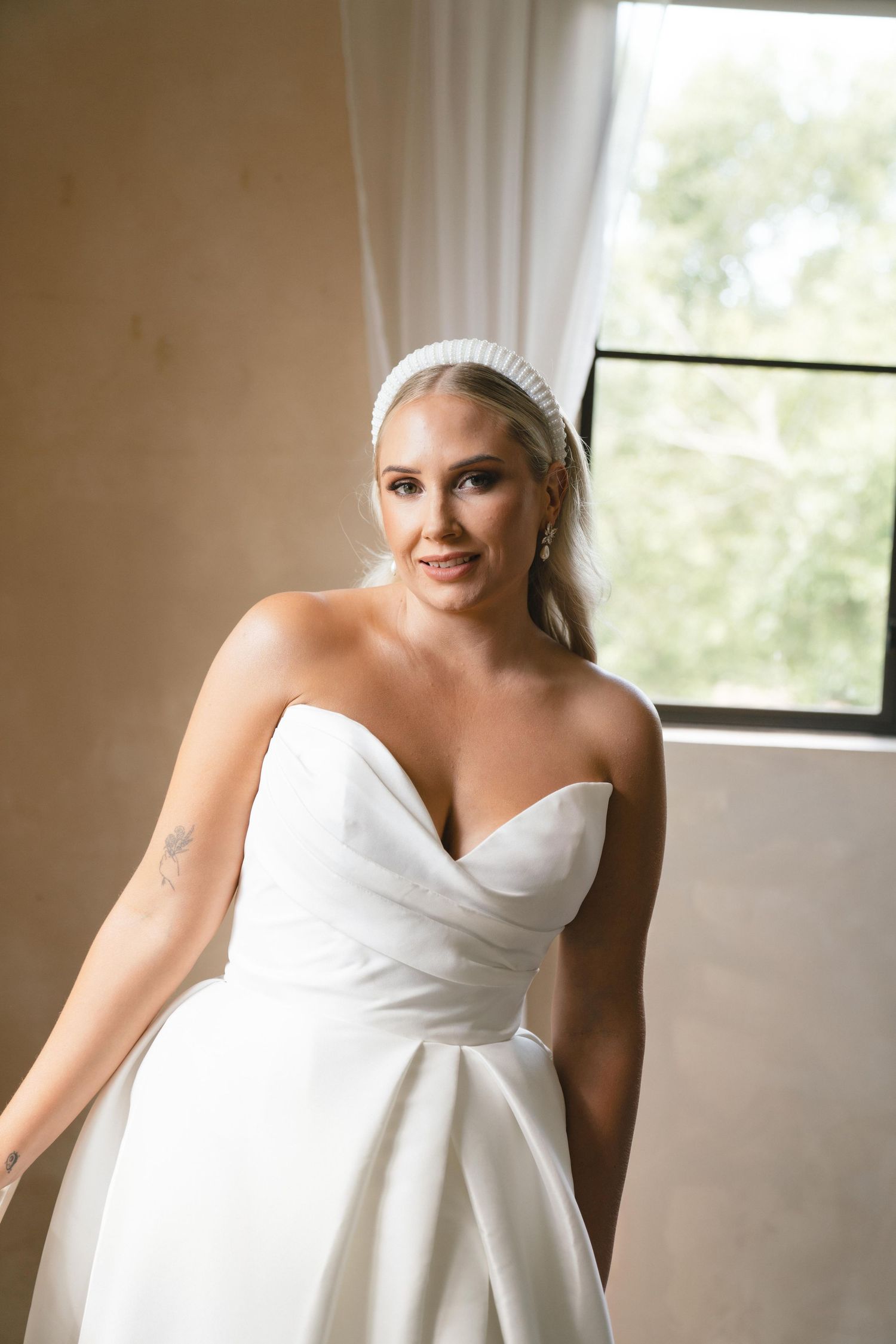 Bride in strapless white wedding gown with sweetheart neckline and pearl headband poses near window.