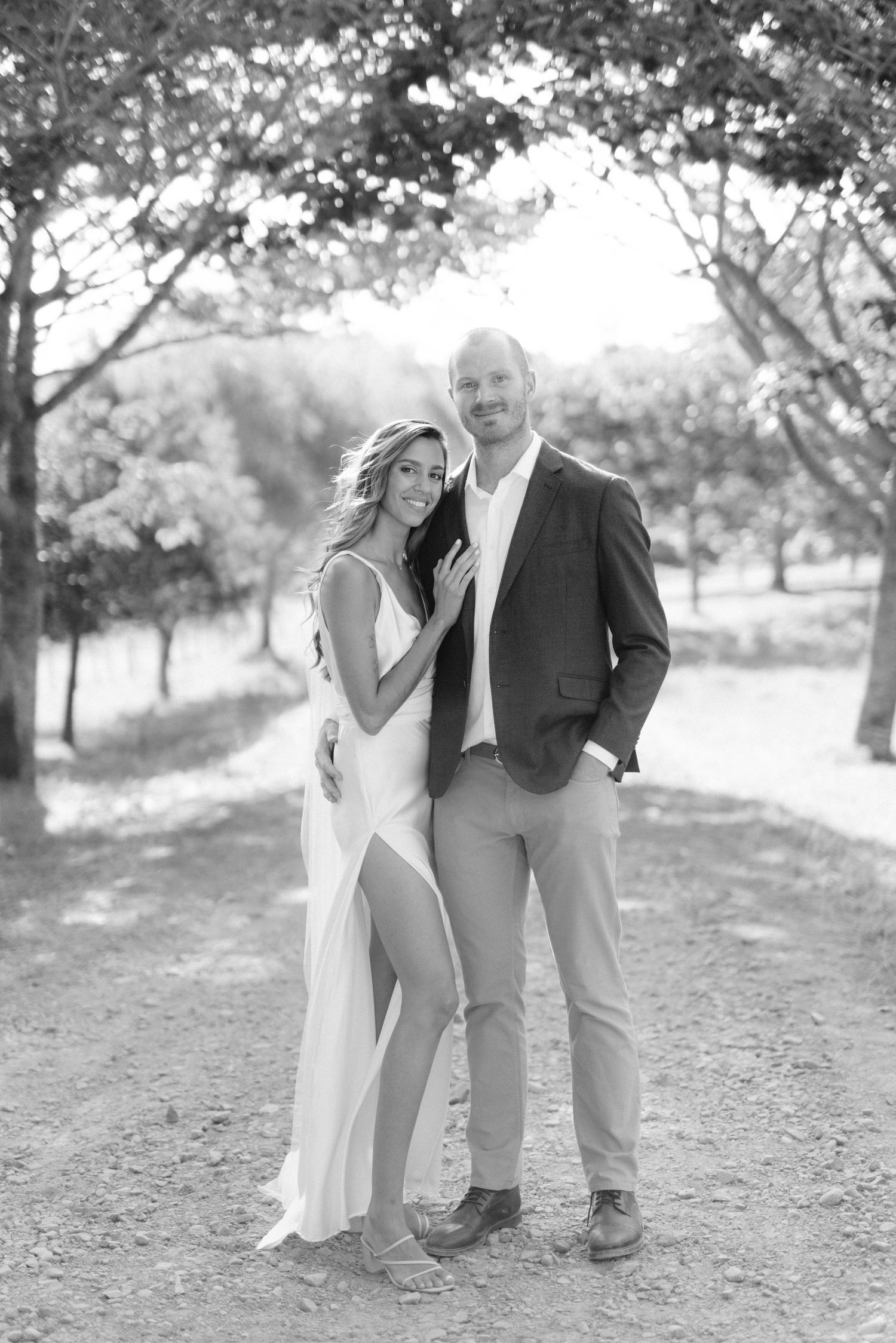 Black and white photo of a couple posing outdoors, woman in white dress and man in blazer, surrounded by trees.