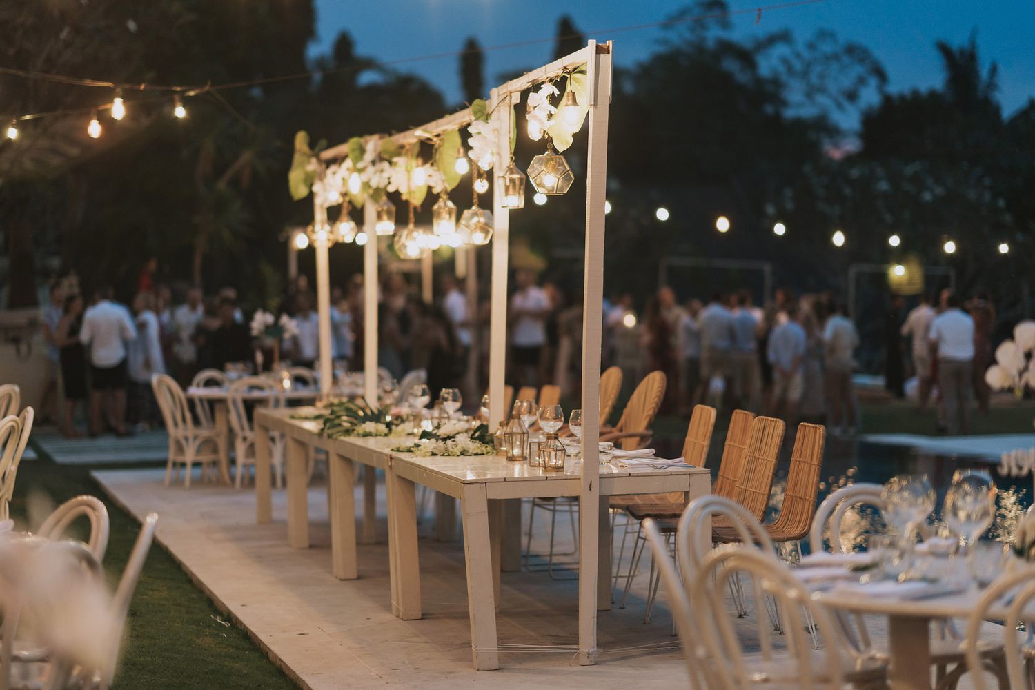 Long dining table with floral arrangements and string lights set for an outdoor wedding celebration.
