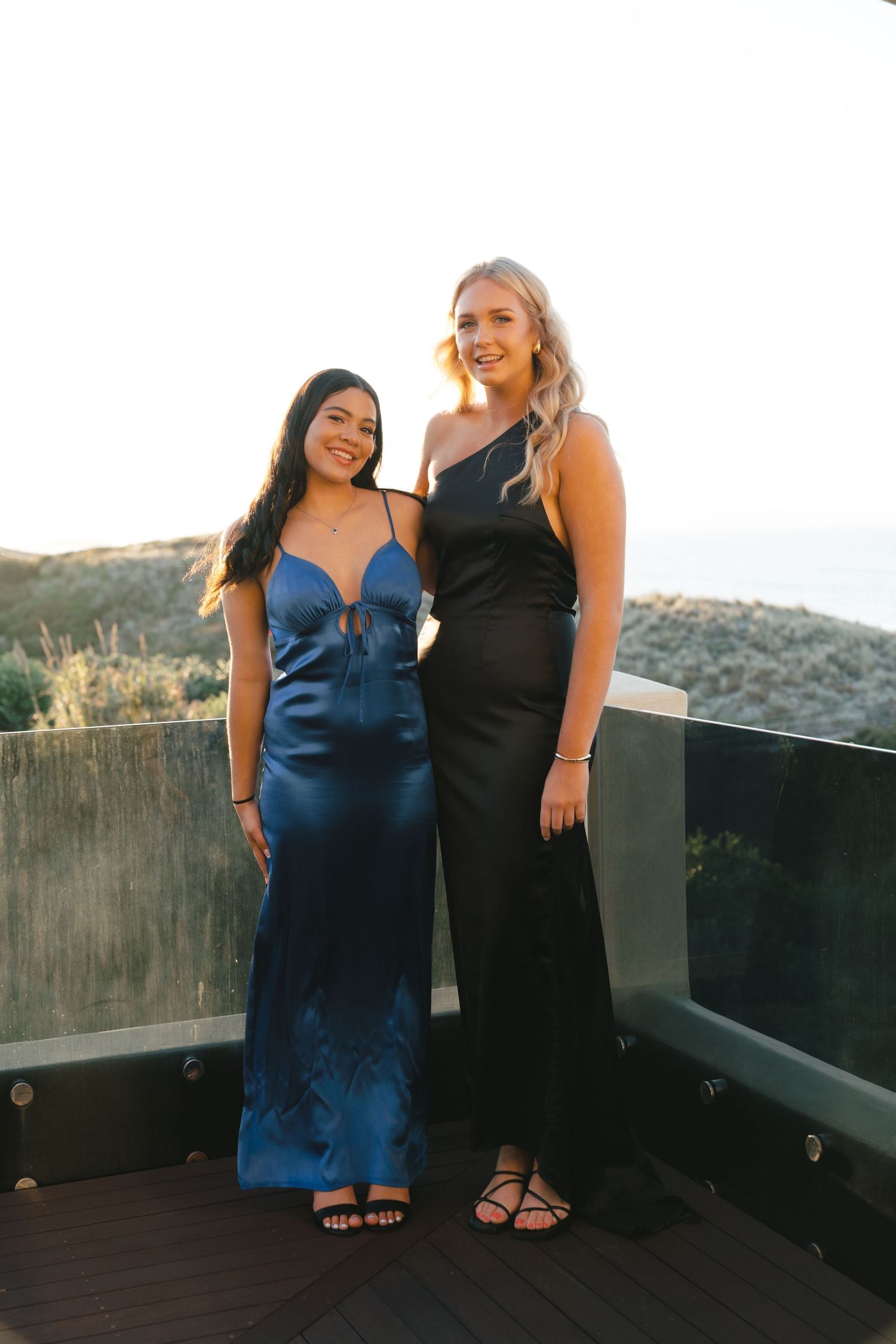 Two friends in contrasting blue and black dresses posing on outdoor deck at sunset.