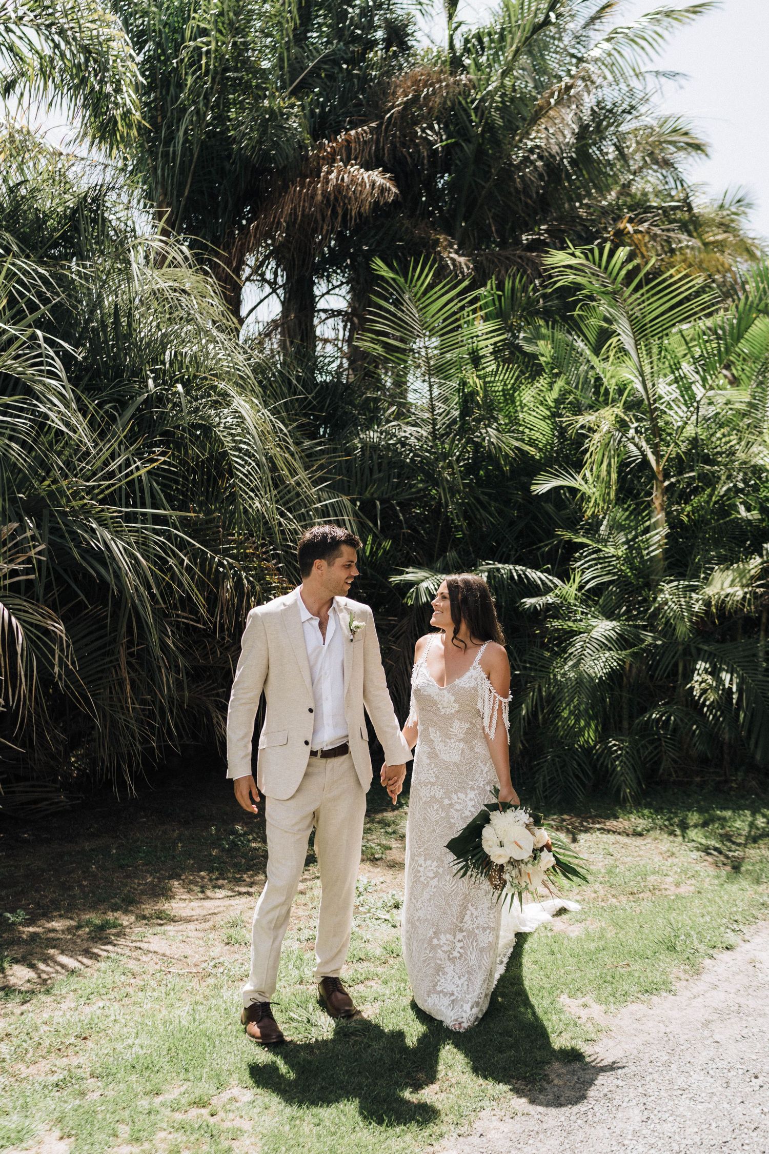 A romantic couple in white wedding attire stroll along a palm-lined tropical path on their special day.