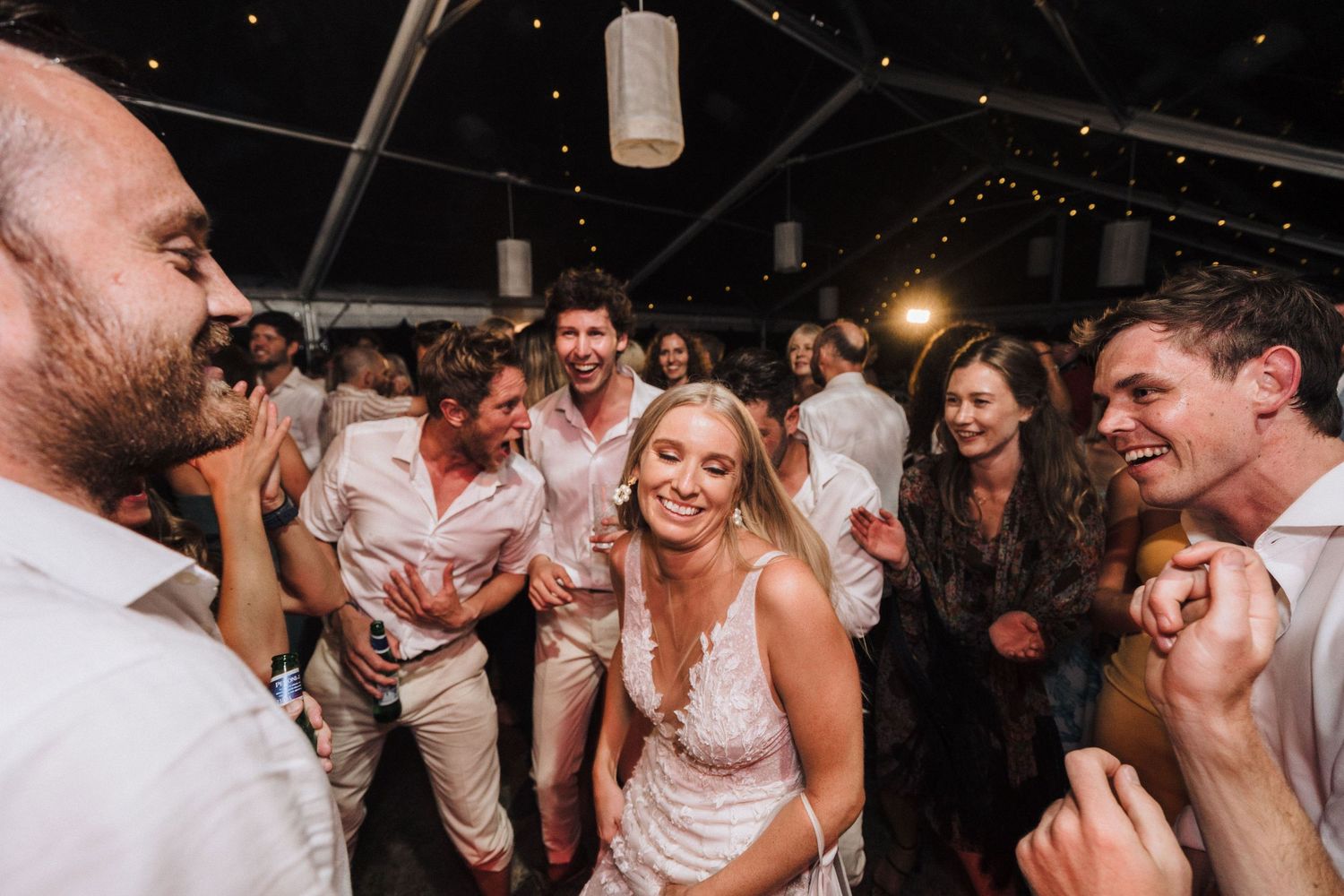 A lively dance party scene under a tent with string lights glowing overhead at an evening celebration.
