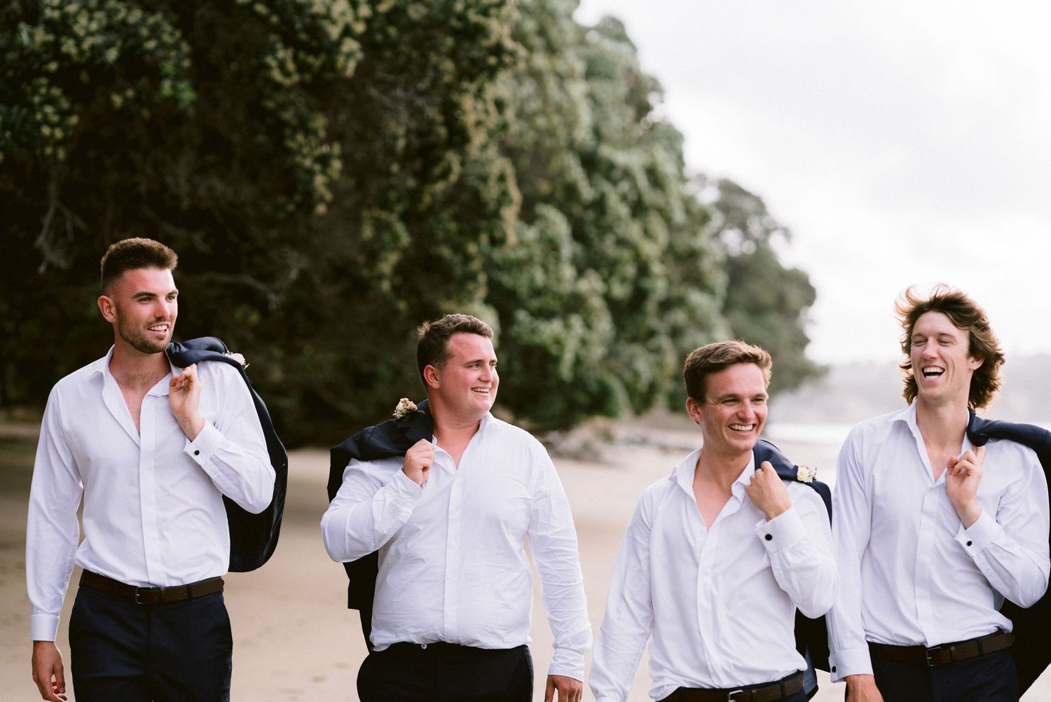 Wedding party members in white shirts and dark jackets laughing together on beach.