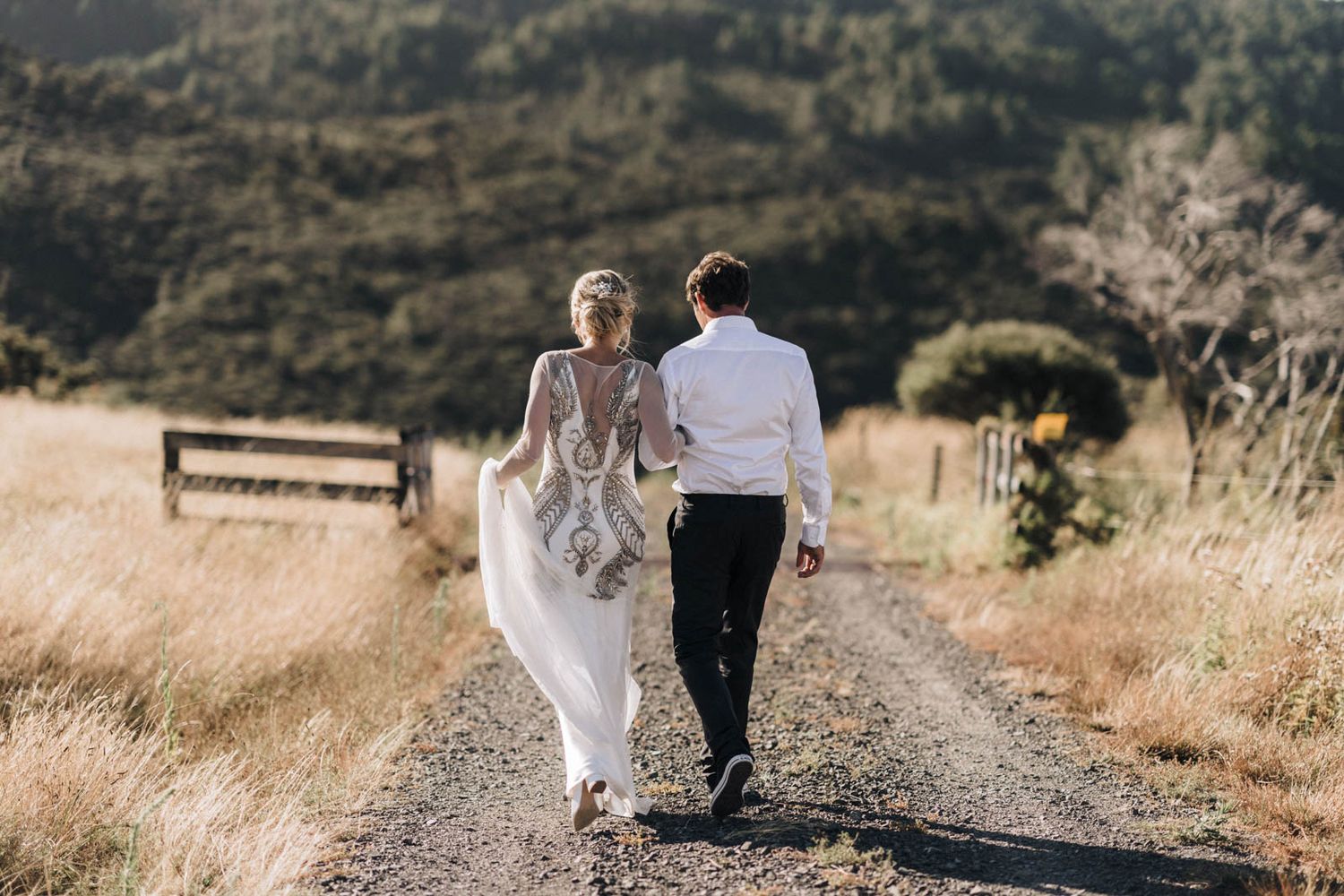 A couple wearing wedding attire walks hand in hand along a rustic path through rolling golden hills at sunset.