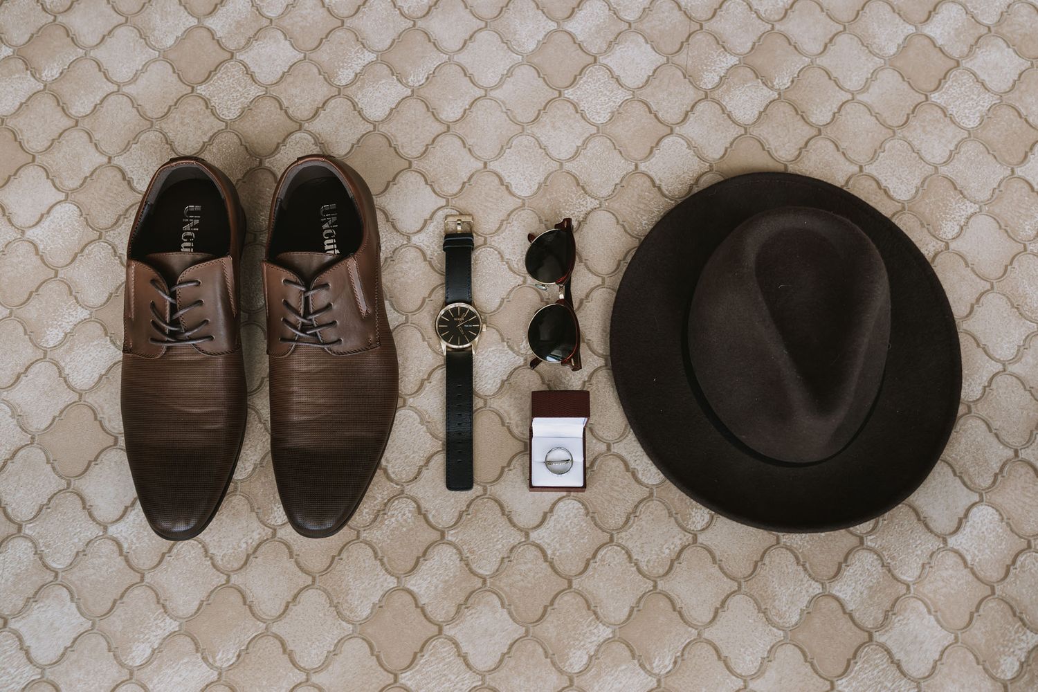 A collection of brown leather shoes, boots, and accessories arranged in a row on a patterned floor tile surface.