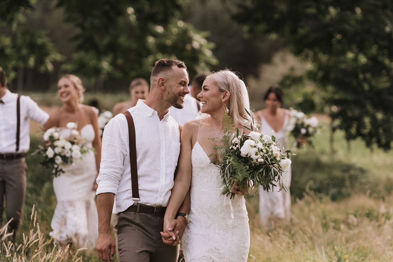 Wedding party enjoying candid moment in rustic outdoor setting with white bouquets and casual attire.
