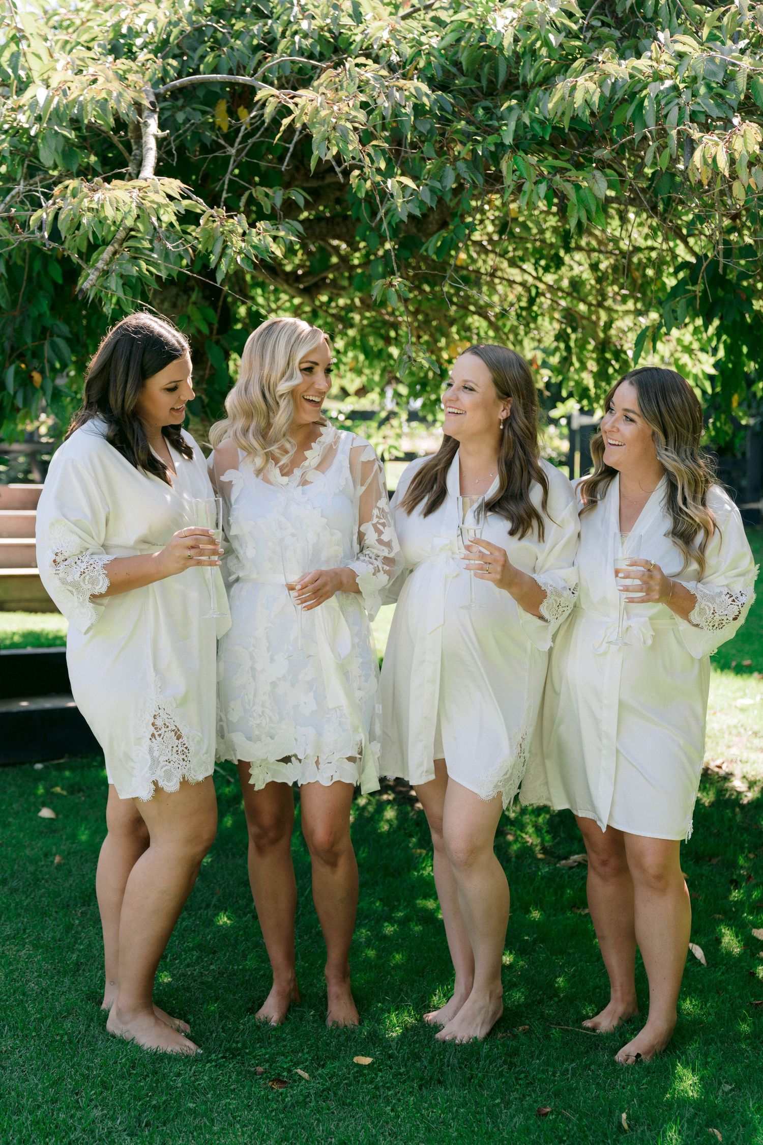 Four friends in white robes and lace dresses stand together barefoot on grass during a special outdoor celebration.