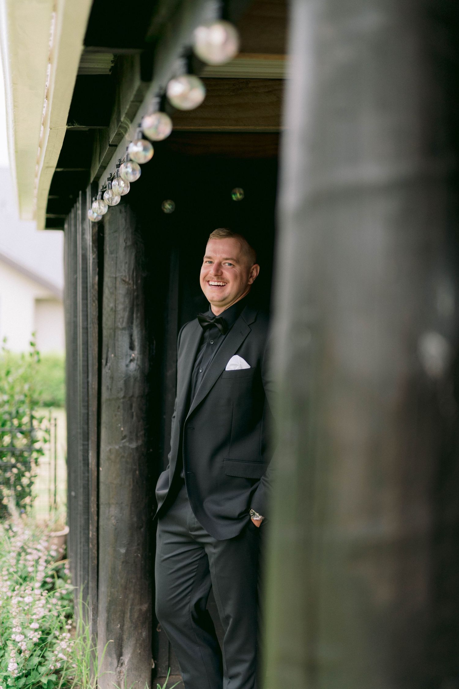 A groom poses in a black tuxedo against a rustic wooden pergola creating artistic pre-wedding portraits.