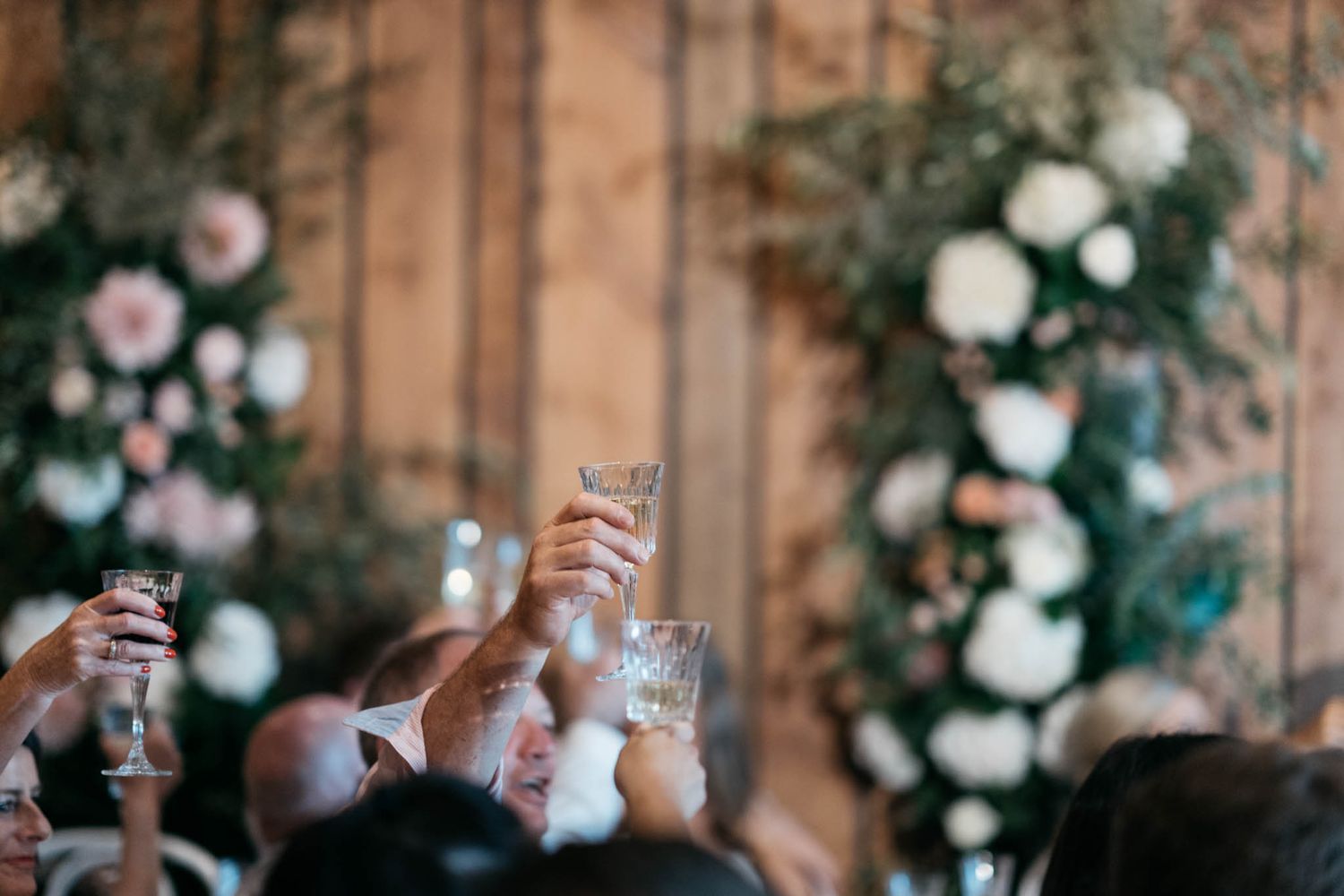 Hands raise champagne glasses in a toast during a celebration with white floral decorations and wooden walls in background.