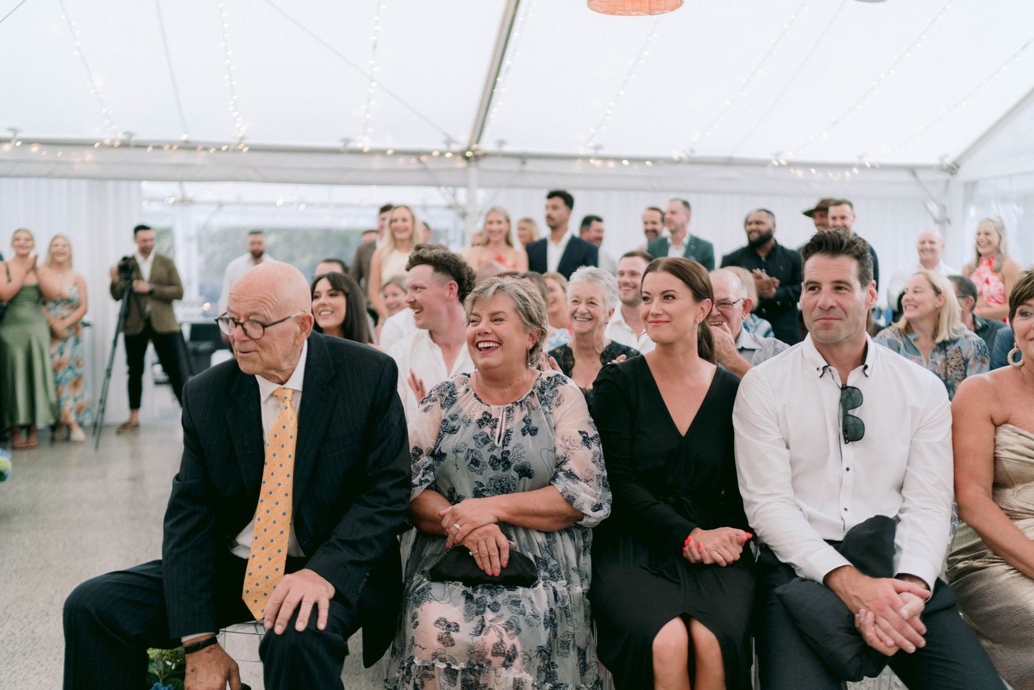 Wedding guests seated under a white tent during an outdoor ceremony.