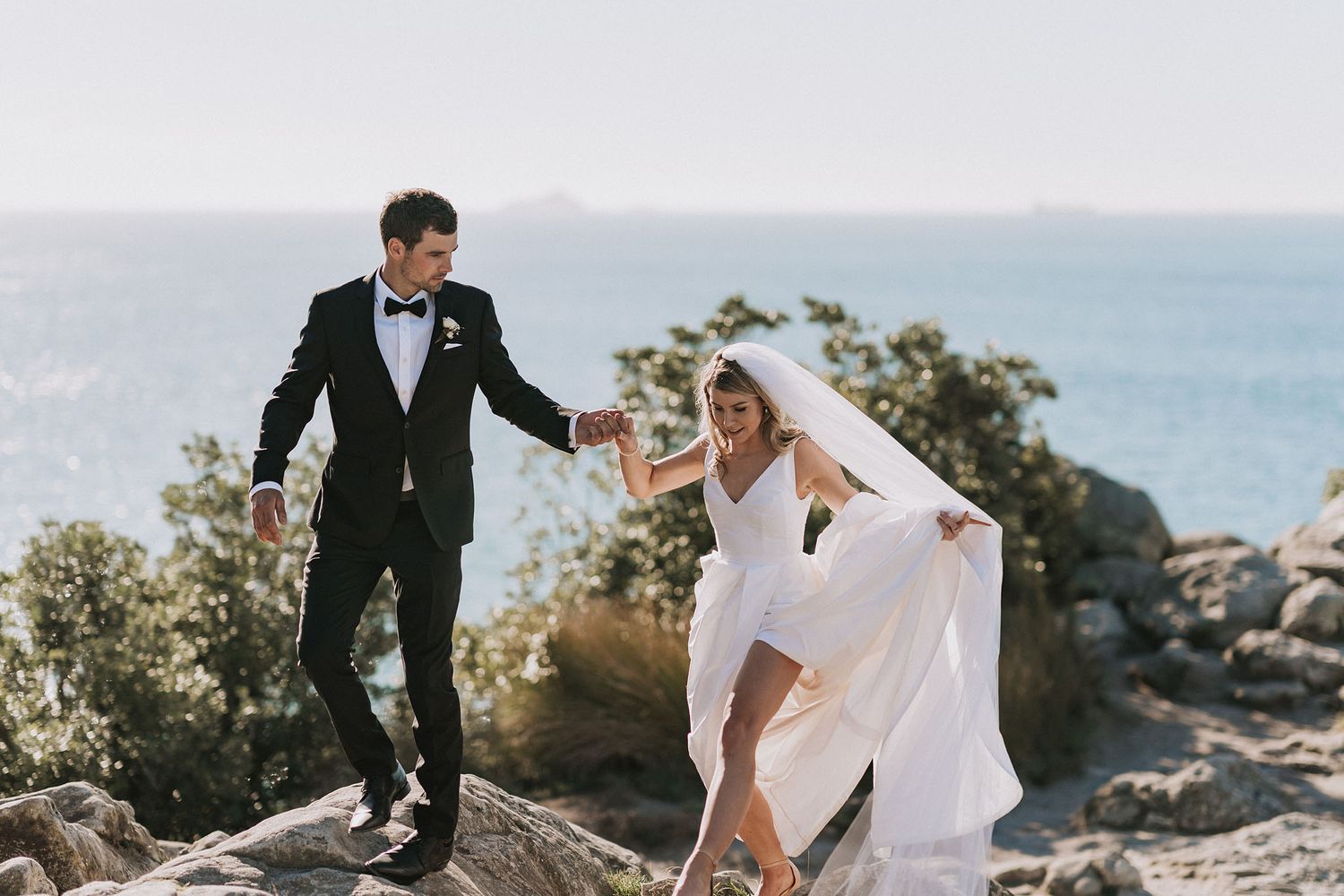 Newlywed couple walks hand in hand along rocky coastal cliffs with ocean views and natural light.