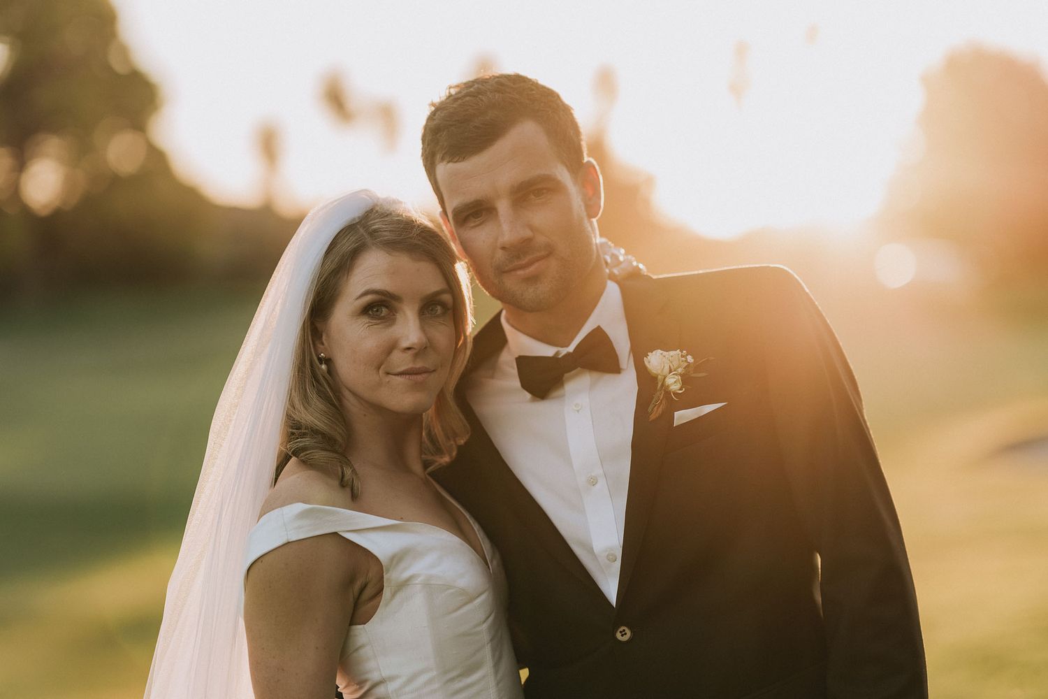Couple in formal wedding attire poses romantically in a garden setting during golden hour sunset.