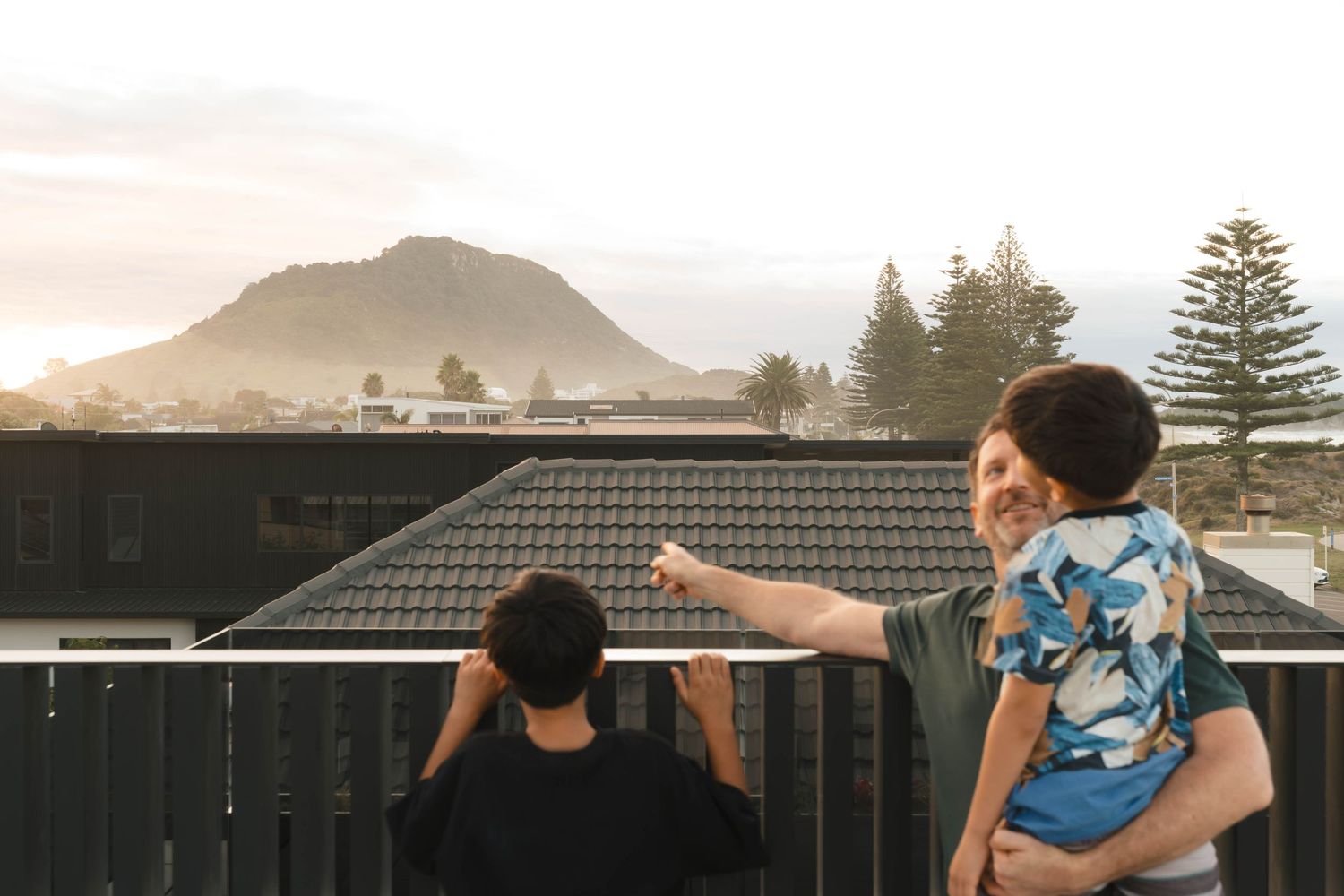 Two people standing on a rooftop deck at sunset with a mountain and Norfolk pines silhouetted in the background.