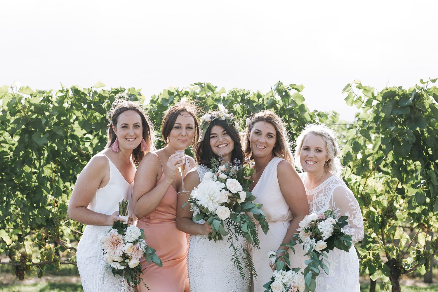 Bridesmaids in peach-colored dresses pose with white floral bouquets in a sun-drenched vineyard setting.