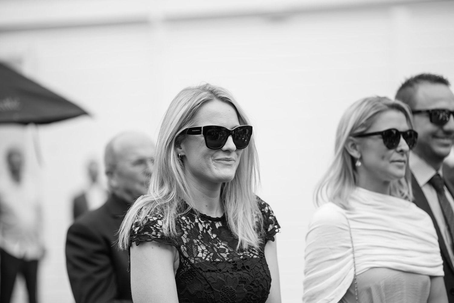 A row of people wearing sunglasses sit together and smile while watching an outdoor event.
